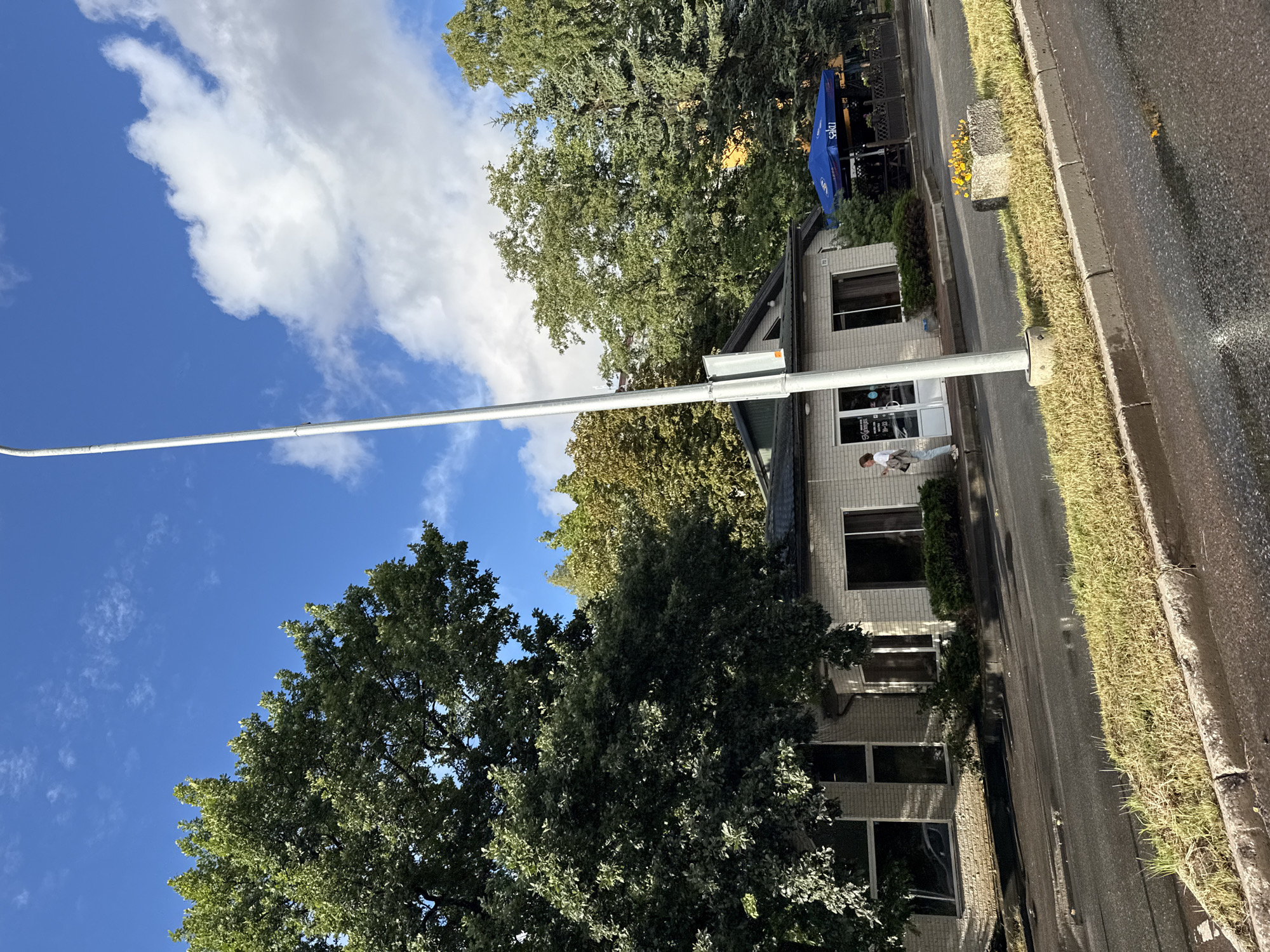 This is a street-level, outdoor, eye-level shot captured on a sunny day in Tallinn, Estonia. The image is oriented vertically, with the sky at the top and the road at the bottom. In the foreground, a light-colored pole extends from the bottom to the top of the frame, suggesting it might be a street light or a traffic signal pole.
The scene features a modern, two-story building with a light brick facade and numerous dark-tinted windows. A person, casually dressed in light-colored pants and a white top, is walking away from the building towards the right side of the image. The building appears to be a commercial establishment, with signage visible on its front entrance. One sign displays the word "Lounge" and operating hours, "10-22".
A bright blue awning or umbrella, also bearing the word "Lounge", is visible over an outdoor seating area to the right of the building. Lush green trees, some with dense foliage, surround the building and line the street, indicating a well-treed urban environment. The street itself is asphalt, and to the right of the sidewalk, there is a strip of dry, yellowed grass. The sky is a vibrant blue, dotted with a few white, fluffy clouds. The lighting suggests it is daytime, likely late morning or afternoon, given the bright sun and shadows.