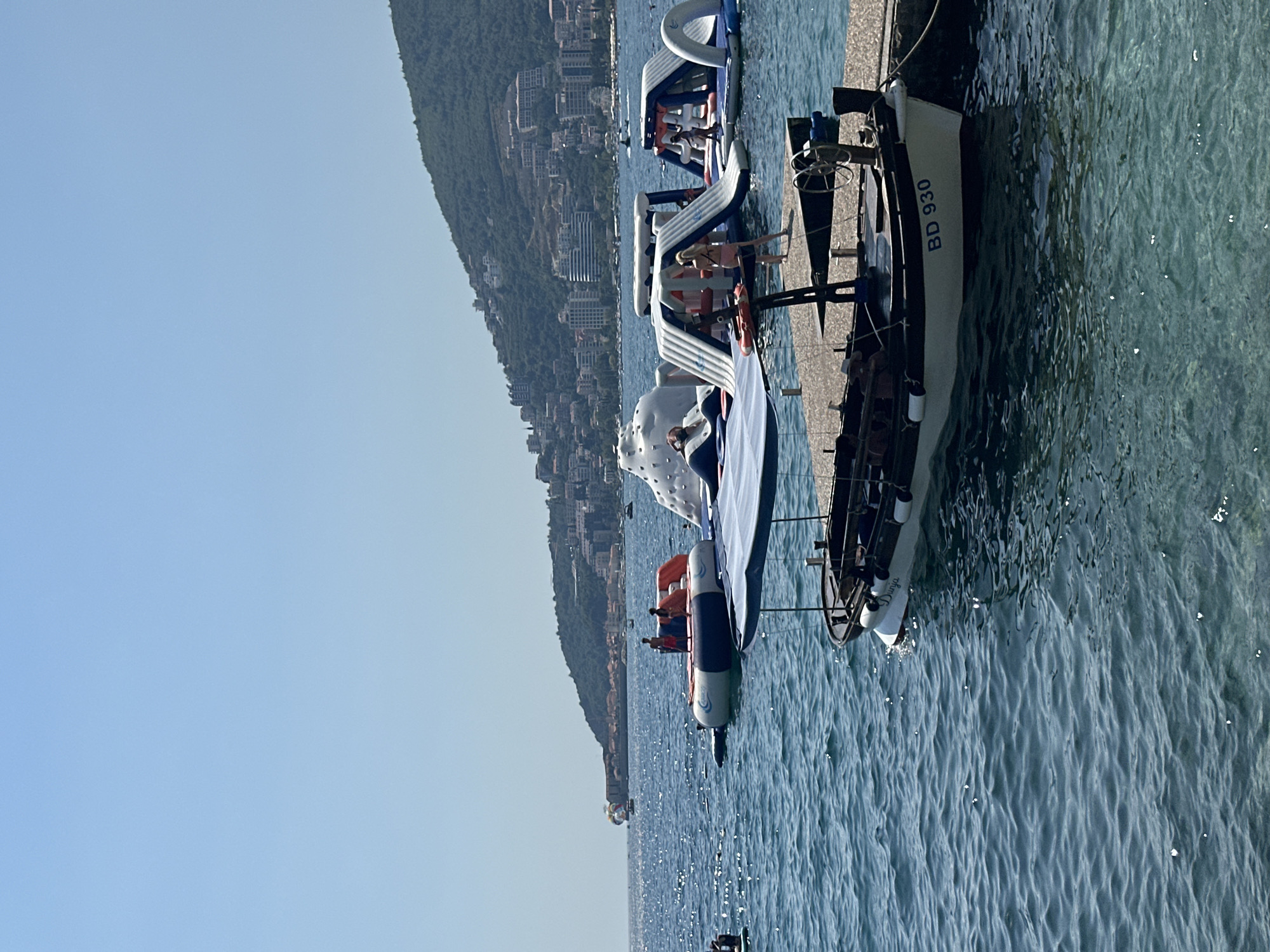 The image depicts a sunny day at an aquatic park in Budva, Montenegro. In the foreground, a small white boat with a blue canopy is docked at a pier, displaying the registration number "BD 930". The boat has a wooden interior and a steering wheel. Beyond the boat, the blue sea is dotted with several inflatable water structures, including slides and trampolines. People are visible on and around these inflatables, enjoying recreational activities. In the background, a lush green hill rises, covered with buildings and trees, characteristic of the Budva coastline. The clear blue sky suggests fair weather.