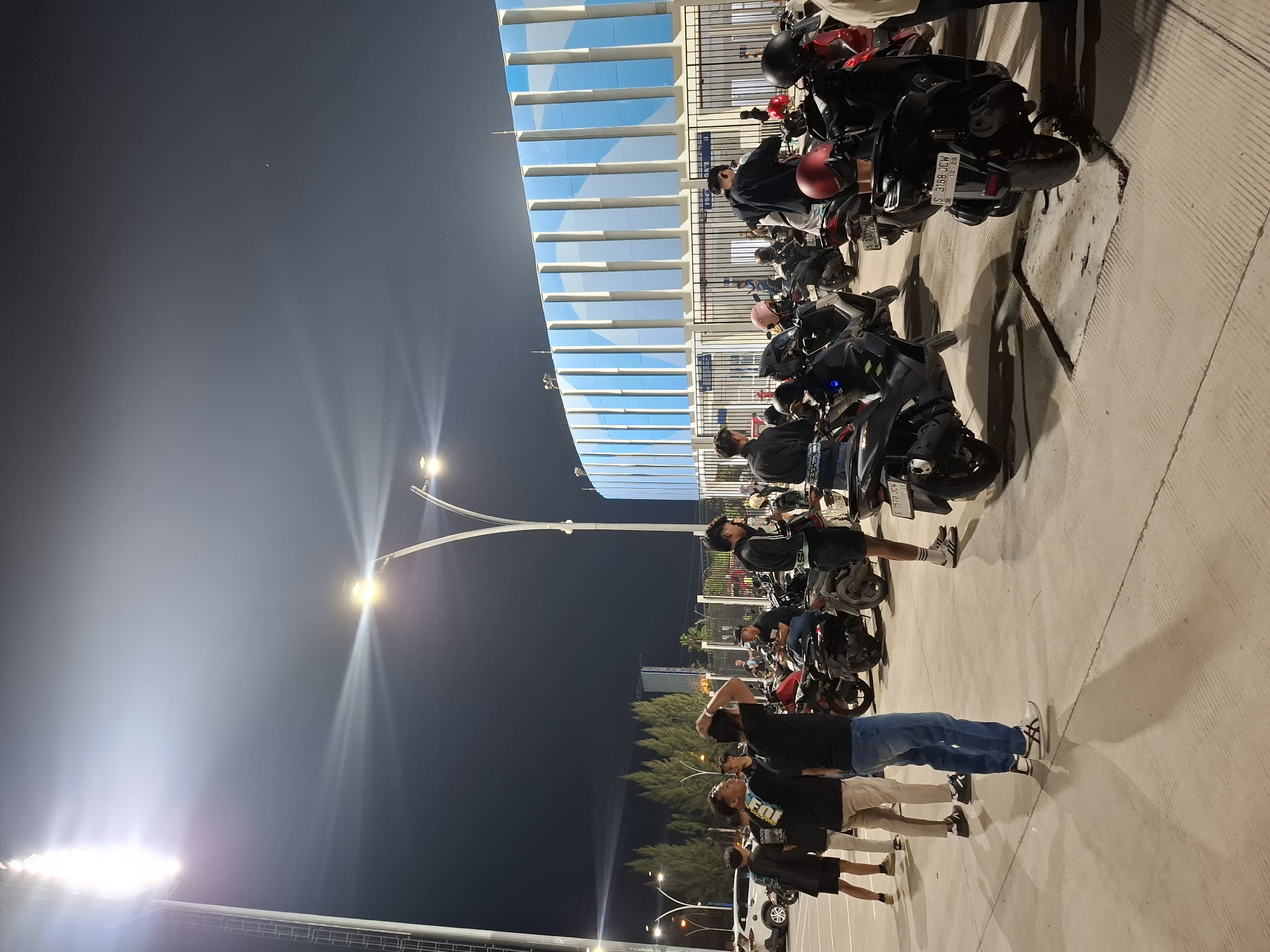 The image depicts an outdoor scene at night, illuminated by bright stadium lights. A group of people, predominantly young men, are gathered in an open area. Many of them are standing near a collection of parked motorcycles. The environment appears to be a public space, possibly a parking lot or an area adjacent to a large building. In the background, a modern building with a facade of blue and white vertical panels is visible. The ground is paved with concrete, showing visible seams. The sky is dark, indicating it is nighttime. The overall atmosphere suggests a gathering related to an event, perhaps a sporting match or concert, given the stadium lighting and the presence of many people and vehicles. The location is identified as Perumda Deket, Indonesia.