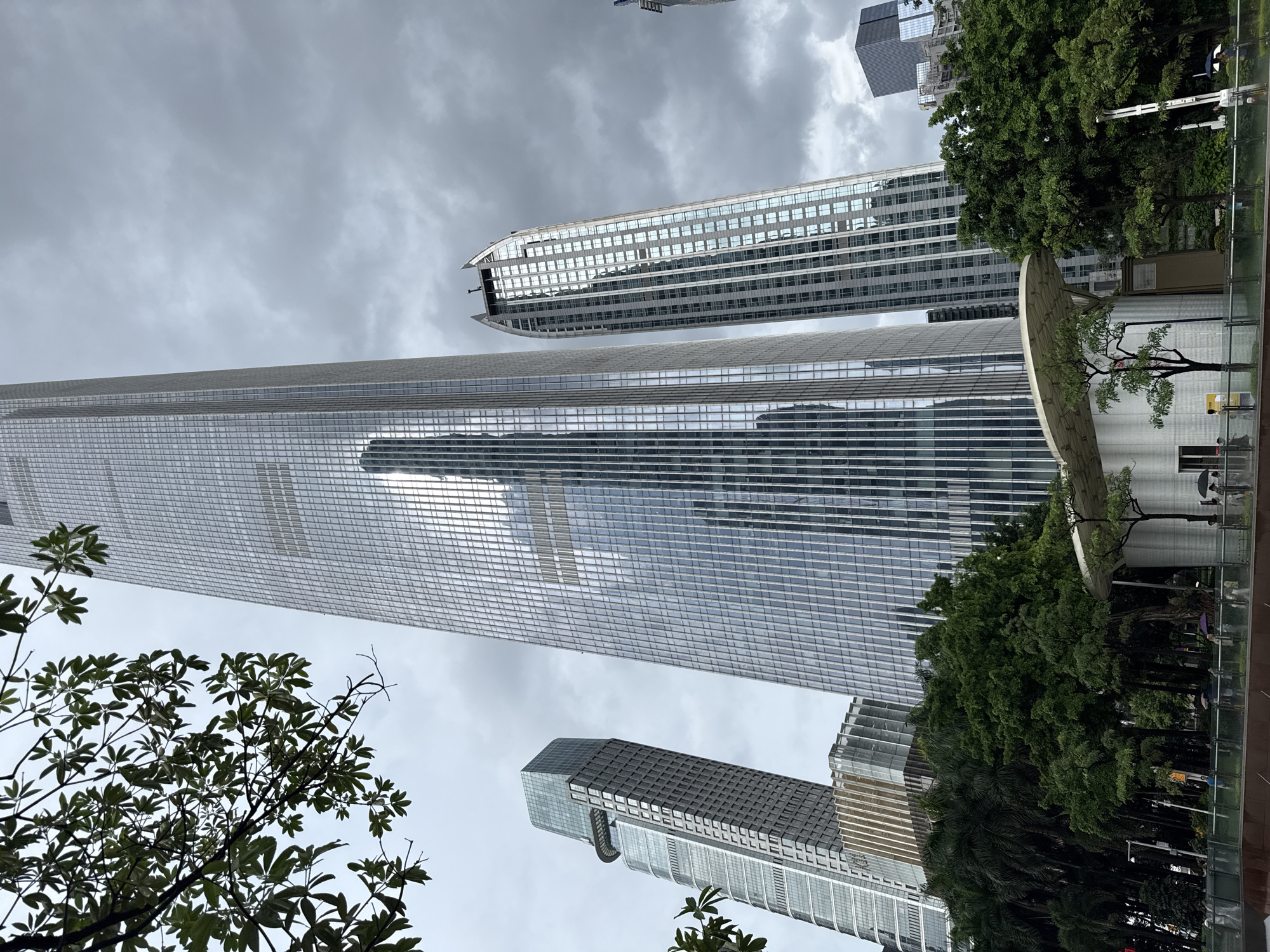 The image shows a view of modern skyscrapers in Tianhe District, China, under a cloudy sky. In the foreground, there is a street with people walking and holding umbrellas, suggesting it might be raining. Lush green trees and a white, modern building structure are also visible in the foreground. The skyscrapers are made of glass and steel, reflecting the sky and surrounding buildings. The tallest skyscraper dominates the center of the image, with other buildings of varying heights and architectural styles to its left and right. The overall scene depicts an urban environment with a mix of nature and architecture.