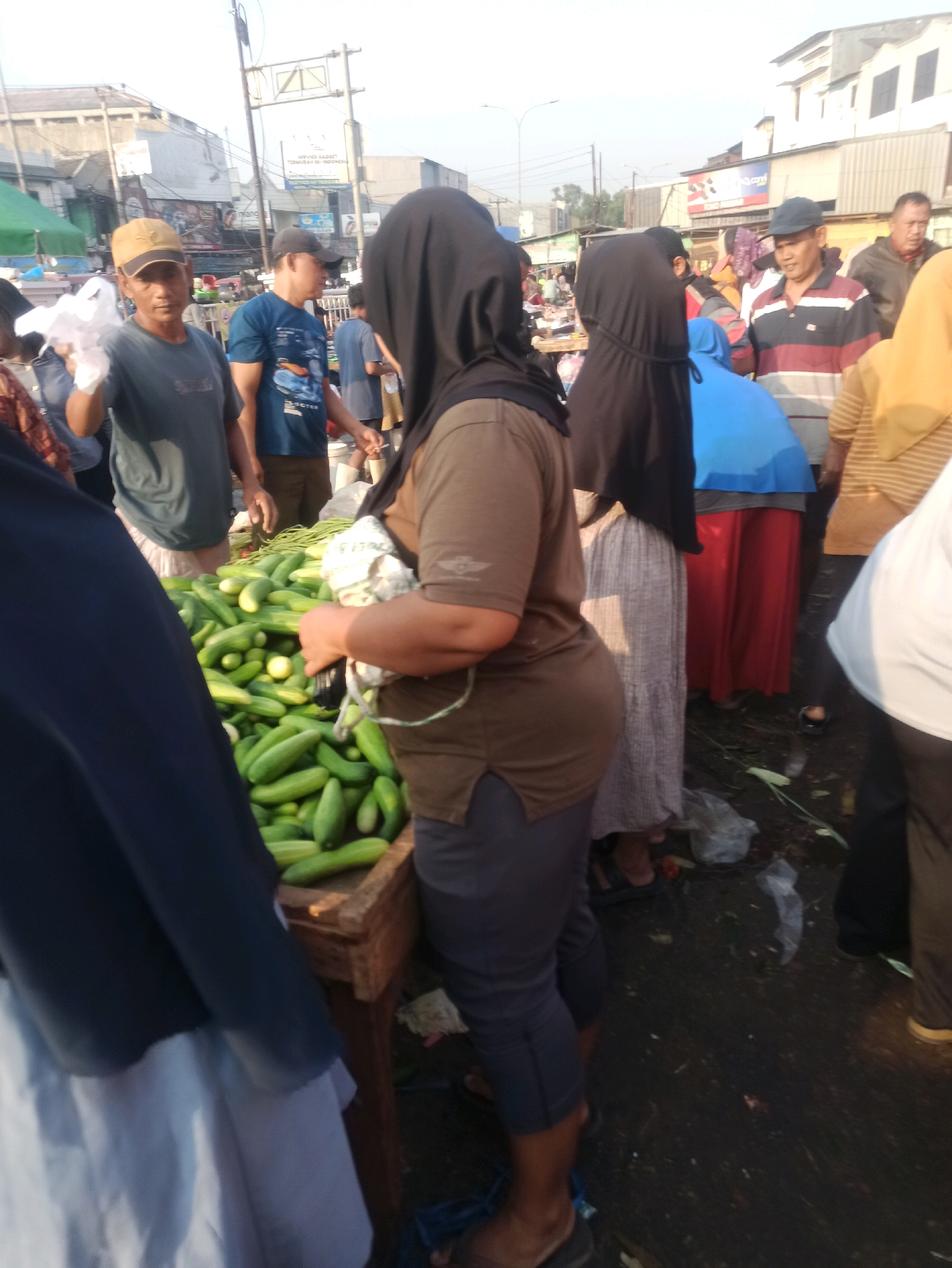 The image depicts a street market scene in Cikarang, Indonesia. Several individuals are present, some appearing to be vendors and others customers. In the foreground, a large pile of cucumbers is displayed on a wooden stand. A woman wearing a black headscarf and a brown shirt is standing behind the cucumbers, holding a white plastic bag. To her left, a man wearing a blue t-shirt and a cap is holding a plastic bag. Another man, also wearing a cap, is standing further back, dressed in a striped shirt. The background shows buildings and overhead wires, indicative of an urban environment. The ground is paved and appears somewhat damp, with some debris scattered around. The lighting suggests it is daytime, possibly late afternoon.