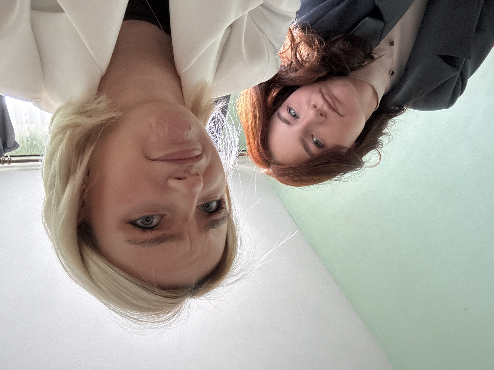Two women are featured in a low-angle portrait, looking down directly at the camera, which appears to be placed below them. The image is oriented with the subjects' heads pointing towards the bottom.
On the left side of the frame (when viewed correctly), a woman with short blonde hair and light blue eyes wears a white blazer or jacket. Her expression is somewhat serious or contemplative, with her eyebrows slightly furrowed. To her right, another woman with long, wavy auburn hair and blue-green eyes is visible. She is dressed in a dark blue or black jacket over a light tan or cream-colored top, and a necklace adorned with black clover-shaped charms can be seen around her neck. She displays a subtle, pleasant smile.
The background is simple, consisting of a plain wall, predominantly white on the left side and transitioning to a light mint green on the right. A blurred window or light source is partially visible on the far left, suggesting an indoor setting. The lighting indicates it is daytime. The photograph appears to be a casual selfie or a picture taken from a low vantage point. There is no visible text in the image. While the location context of Sinyavino, Russia, is provided, there are no specific visual cues within the image itself to confirm this.