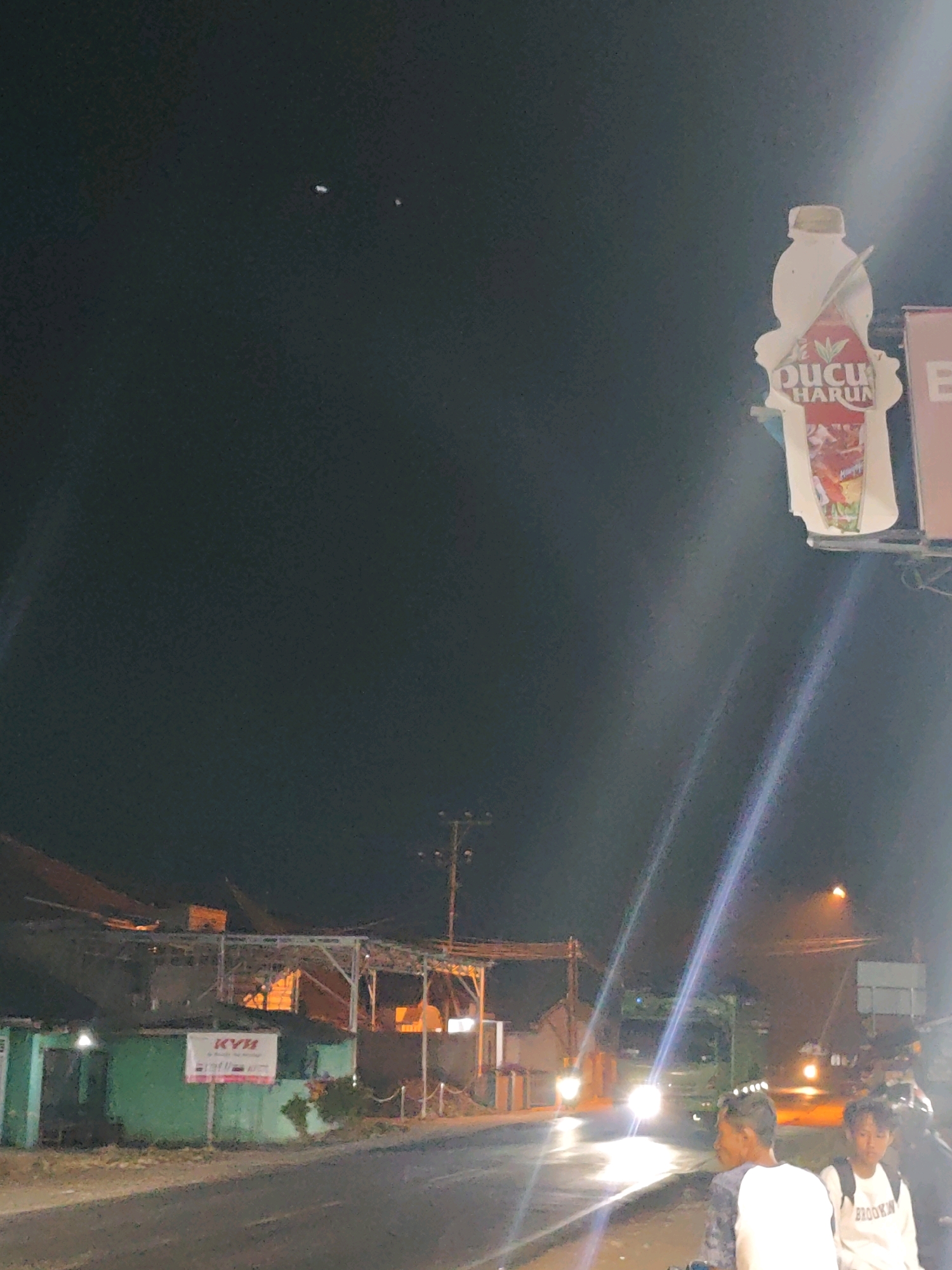 This is a nighttime photograph of a street scene in Gumuk Mas, Indonesia. Two young men are visible in the foreground, looking away from the camera. One is wearing a white t-shirt with "BROOKLYN" written on it, and the other is wearing a patterned t-shirt. Behind them, a street is illuminated by car headlights, suggesting that traffic is present.
In the background, there are buildings with some lights on, and a large advertisement shaped like a bottle is hanging from a pole. The advertisement features the text "Ducu Harun". Further down the street, another sign with the text "KYB" is visible on a green building. The sky is dark, indicating it is nighttime. The lighting in the image is dramatic, with strong lens flares from bright light sources, creating streaks of light across the frame. The overall atmosphere is that of a casual evening street scene.