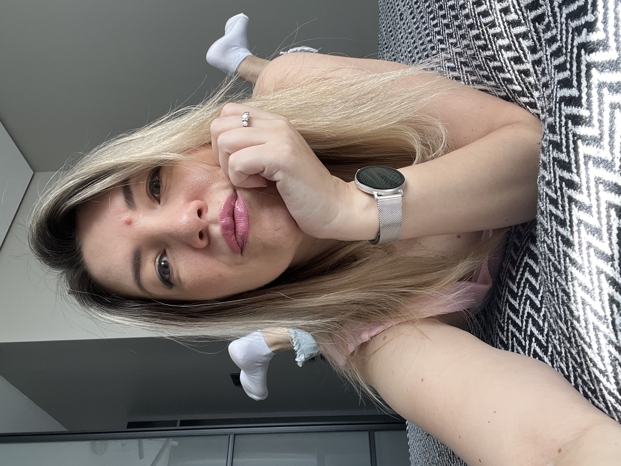 The image shows a woman lying on a bed, with her face propped on her hand. She has blonde hair, pink lipstick, and is wearing a pink top, denim shorts, white socks, and a silver watch. Her feet are in the air behind her. The bed has a black and white patterned blanket. The background shows a closet and a gray wall. The woman appears to be in a bedroom.