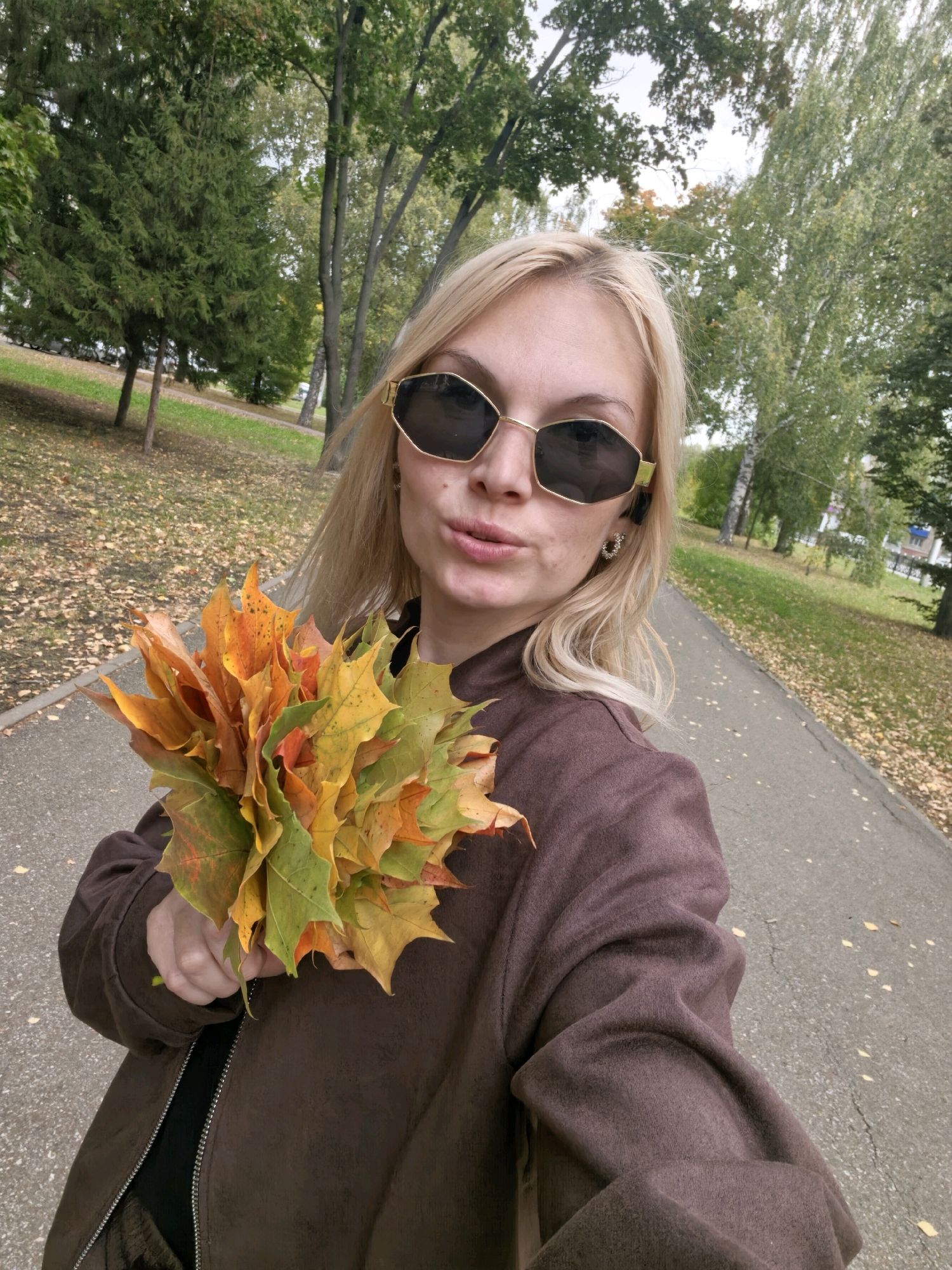 A Caucasian woman with blonde hair and sunglasses is standing on a paved path in a park, holding a bouquet of autumn leaves. She is wearing a brown jacket. The ground is covered with fallen leaves, indicating it is autumn. Trees with green and yellow leaves are visible in the background, as well as a building in the distance. The lighting suggests it is daytime. This scene is consistent with a park environment in Sterlitamak, Russia, during the fall season.