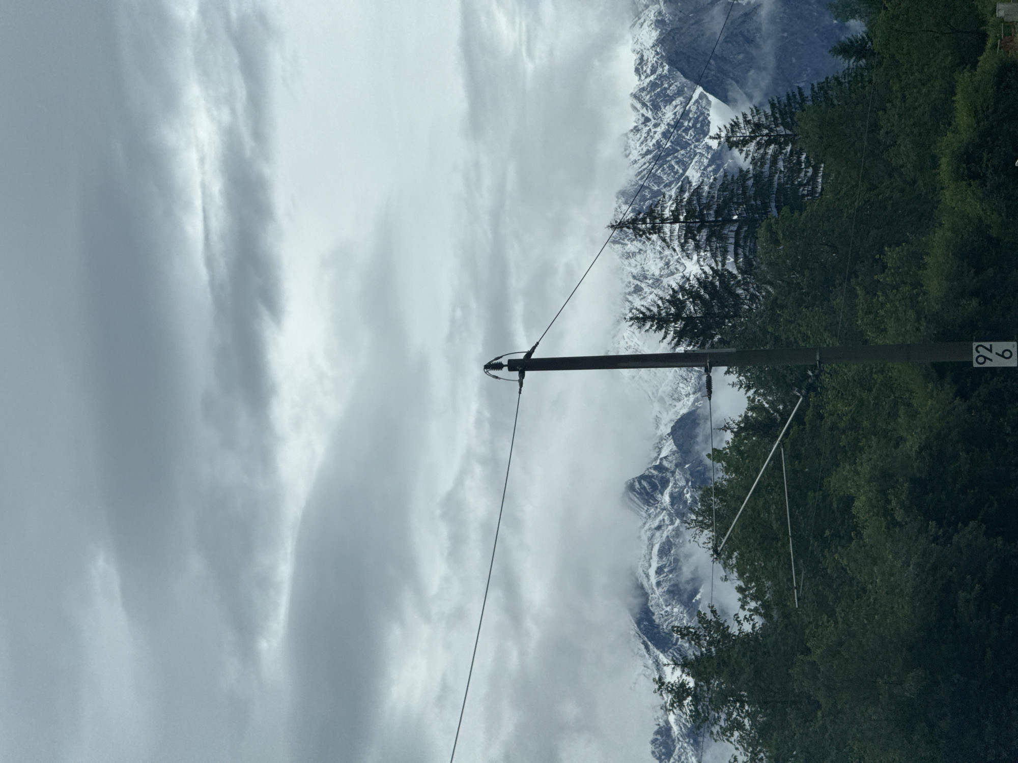 The image, correctly oriented, depicts a majestic mountainous landscape under an overcast sky in Oberau, Germany. In the immediate foreground, a dark utility pole stands vertically on the right side of the frame, featuring insulators and wires that stretch horizontally across the midground. A small, white rectangular sign with the black numbers "92" stacked above "6" is affixed to the pole.
Behind the utility pole and extending across the lower-right portion of the scene is a dense expanse of dark green evergreen trees, likely conifers, their foliage contrasting with the lighter sky and distant peaks.
In the background, imposing snow-capped mountains rise dramatically, their rugged forms partially obscured by low-hanging clouds or mist. The white snow on the peaks suggests a cold climate or high altitude. The sky above is entirely overcast with soft, grey and white clouds, lending a diffused and muted light to the scene. There is no specific activity taking place, and the overall mood is one of serene grandeur and natural vastness under cloudy, possibly misty, daytime conditions.