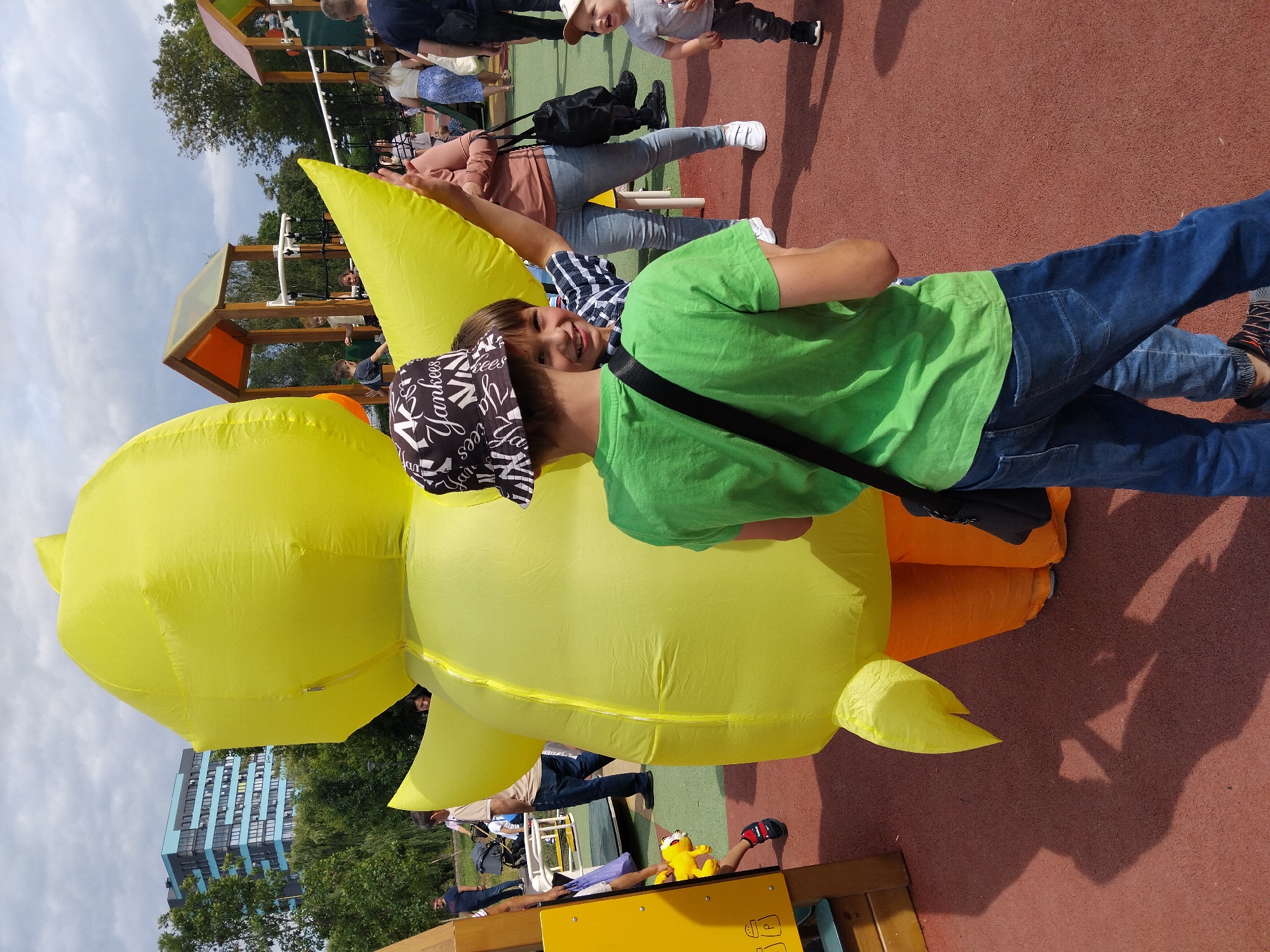 In Rivne, Ukraine, on a cloudy day, a person wearing a large, inflatable yellow duck costume stands on a red rubberized surface, possibly a playground. The duck's head is obscured by the backs of two young boys. The boy in the foreground wears a green t-shirt and a black and white patterned bucket hat. He is looking away from the camera. The boy behind him, to the right, is wearing a blue and white checkered shirt and is looking towards the camera, smiling. Other people are visible in the background, including adults and children, near playground equipment. A modern blue and black building is visible through trees in the distance. The overall scene depicts a public outdoor gathering, likely at a park or recreational area.