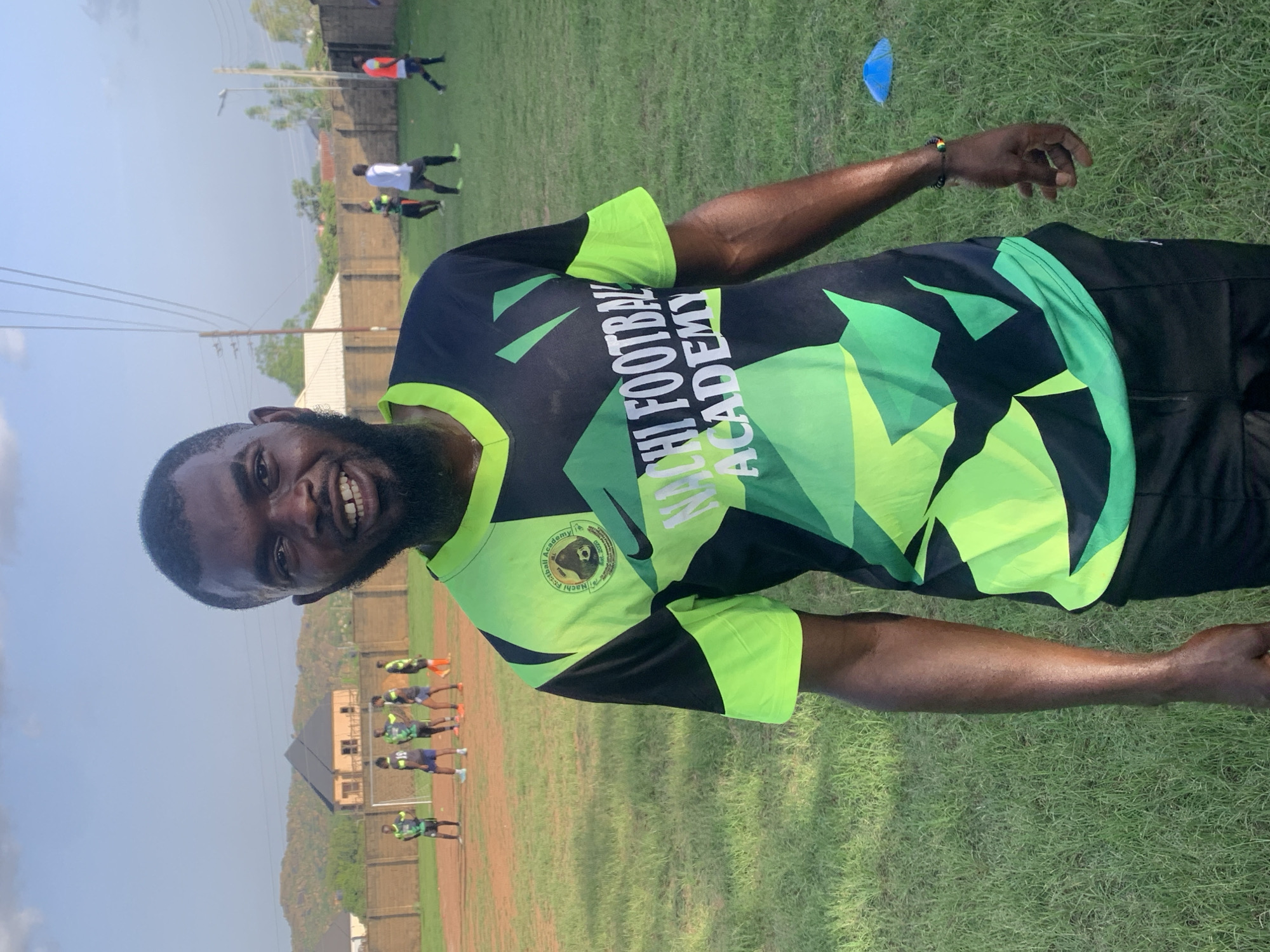 A man with dark skin and a beard stands on a grassy field, smiling at the camera. He is wearing a black and lime green t-shirt with the words "NACHI FOOTBALL ACADEMY" printed in white and green on the front. He is also wearing black shorts and a bracelet on his right wrist. In the background, several other people, presumably football players, are visible on a dirt field with a goalpost. There are buildings and trees in the distance, as well as a fence surrounding the field. The sky is blue with some clouds. The image appears to have been taken in Jos, Nigeria, where Nachi Football Academy is located.