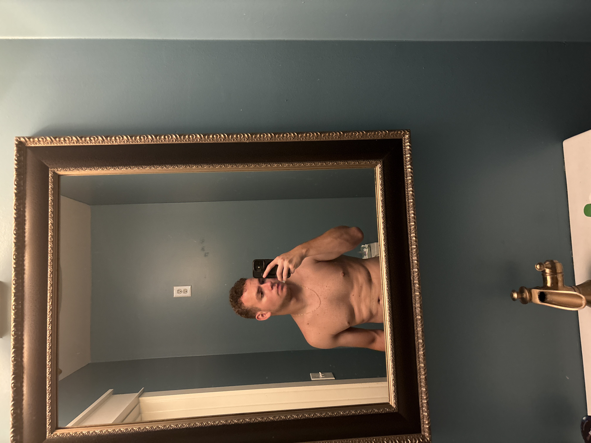 In a bathroom in Panama City Beach, United States of America, a young, fair-skinned man with short brown hair is taking a selfie in a mirror. He is shirtless, wearing a thin gold chain, and holding his phone in his right hand. His left arm is by his side. He appears to be in his early twenties and has a lean, muscular build. The bathroom walls are painted a teal color, and the mirror has an ornate, gold-trimmed frame. Below the mirror, a golden faucet with a unique, angular design is visible on a white sink. The overall impression is that of a private space, likely a home bathroom, captured in a casual moment.