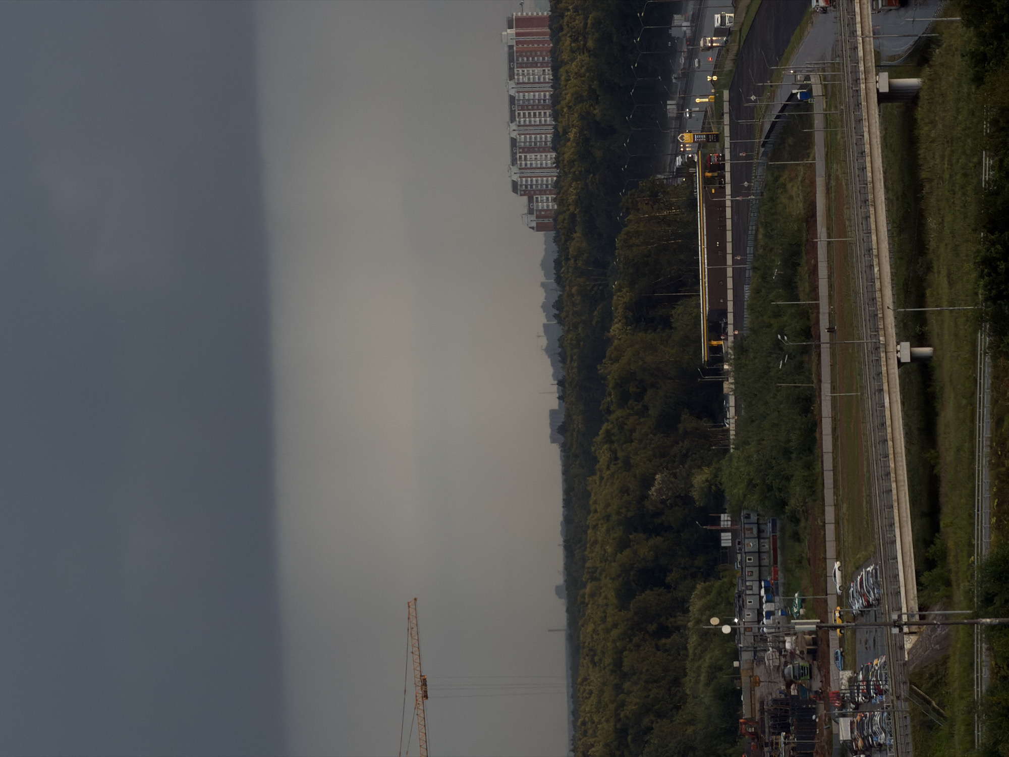 The image depicts a hazy sky over a semi-urban landscape. In the foreground, a multi-lane highway is visible, with several vehicles, including trucks and cars, traveling on it. A concrete overpass crosses the highway. To the left of the highway, there is a large parking lot filled with cars and a construction site with portable buildings and construction equipment. A yellow construction crane is visible in the distance. Further in the background, a dense green forest stretches across the landscape, with a few tall buildings of apartment complexes visible above the trees. The lighting suggests it is either early morning or late afternoon, with overcast conditions contributing to the diffused light.