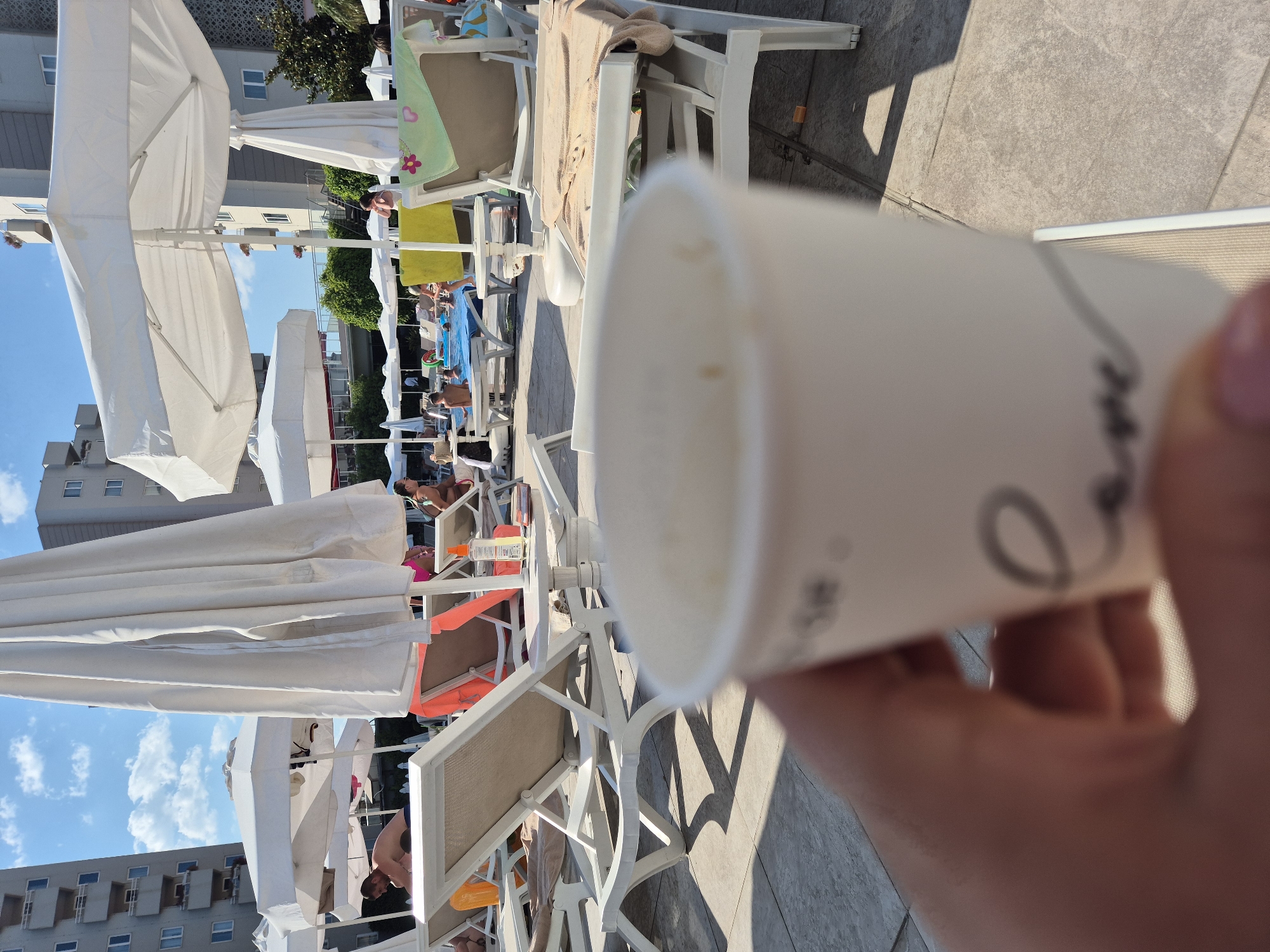 The image captures a sunny day at an outdoor swimming pool area. In the foreground, a hand holds up a white paper cup, obscuring part of the view. The cup appears to have some faint, dark markings on it, possibly a logo or design, though they are not clearly discernible.
The background reveals a scene filled with lounge chairs, many of which are occupied by people relaxing. White umbrellas are spread out, providing shade. Some individuals are in swimwear, suggesting they are either swimming or sunbathing. One person is partially visible in a pool. In the distance, there are multi-story buildings, hinting at a resort or hotel complex. The sky is blue with scattered white clouds, and the sunlight casts shadows on the tiled ground, indicating it is daytime. The overall atmosphere seems relaxed and leisurely, typical of a vacation setting. The crops provided offer closer views of the lounge chairs, umbrellas, and people, as well as a clearer look at the paper cup's markings, though they remain indistinct.