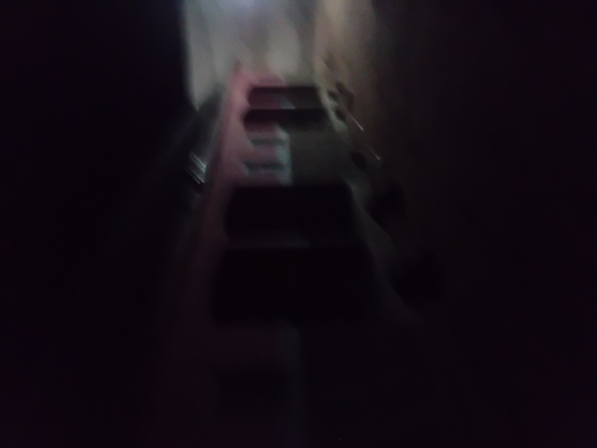 A blurred, night-time photograph depicts the exterior of a building with a red facade. Multiple doorways or openings are visible along the building's front, some appearing as dark rectangular shapes. The ground in front of the building is unpaved and appears to be dirt or gravel. A faint light source illuminates the scene from the right, casting a soft glow on the building and the surrounding area. The image is low in quality due to motion blur and low light conditions, making it difficult to discern specific details or identify any individuals or objects clearly. The location is specified as Jauro Yinu, Nigeria.