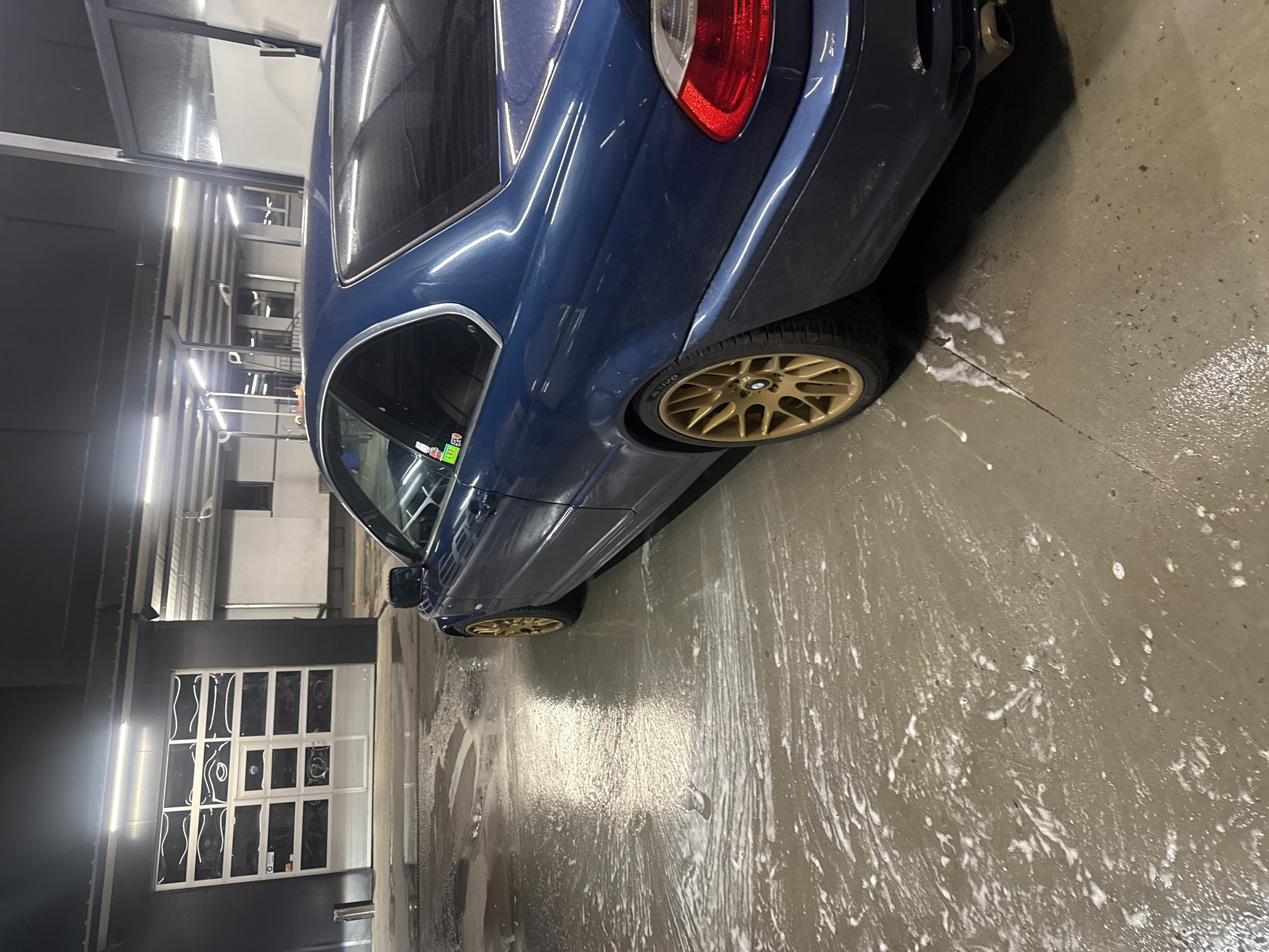 A blue coupe with gold wheels sits in what appears to be an indoor or covered car wash area. The floor is wet with soapy water. The back of the car is in the foreground. In the background, there is a building with windows and what looks like a staircase. The photograph does not depict any specific features recognizable to locate the scene in the city of Burgas, Bulgaria.