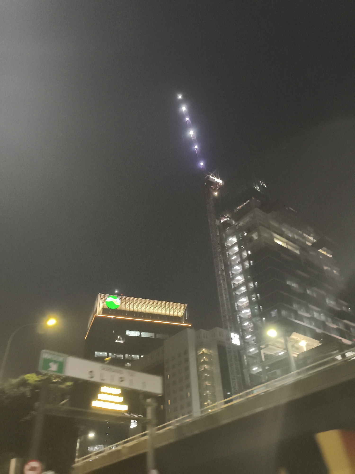 The image captures a nighttime urban scene in West Jakarta, Indonesia, with a focus on a large building under construction, identifiable by a towering crane. The building's structure is illuminated, showing multiple floors and scaffolding. Adjacent to the construction site is another building with a prominent green circular logo featuring a wave-like design. A street lamp with a yellow glow is visible on the left, contributing to the ambient lighting. Below these structures, a sign with text and a green directional icon is mounted on a pole. Further down, an illuminated sign displays yellow block letters. The foreground shows a portion of an elevated road or overpass, with a blurred out-of-focus view suggesting the photo was taken from a moving vehicle. The sky is dark and hazy, indicating nighttime conditions, possibly with mist or light pollution.