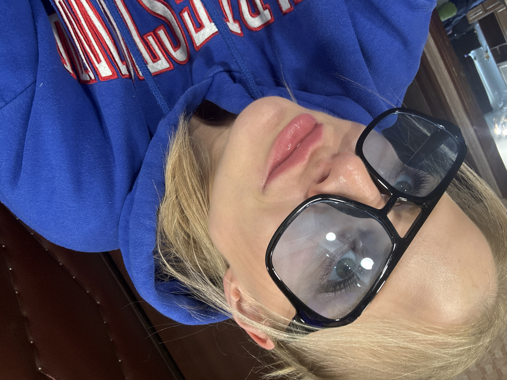 A selfie taken from a slightly angled, upside-down perspective shows a woman with blonde hair, light skin, and glasses. She wears a blue hoodie with white and red lettering. She is indoors and possibly sitting on a brown leather chair or couch. The location in Saint Petersburg, Russia, is not discernible from the image.