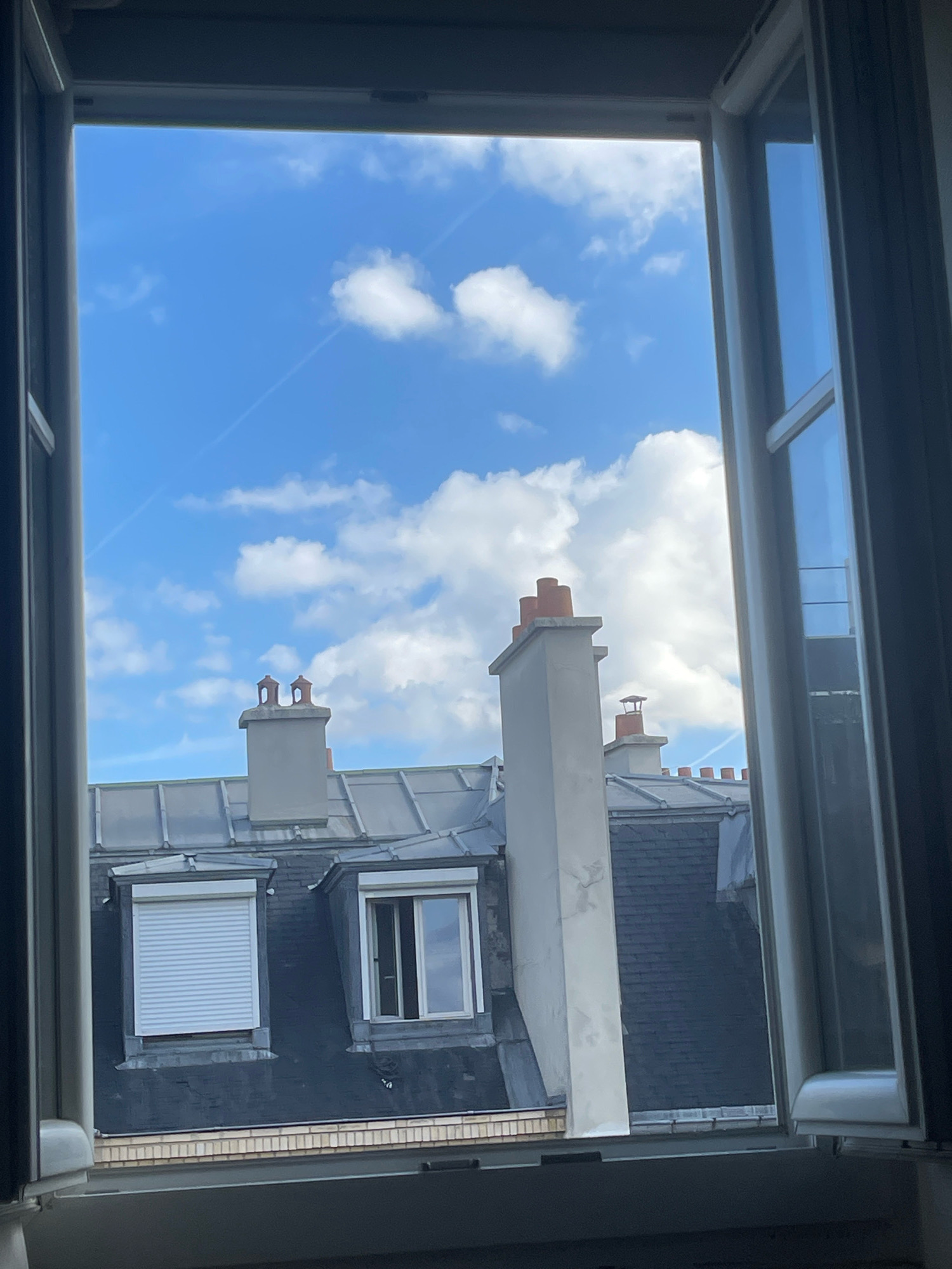 This is a photograph taken from an open window in Paris, France. The view looks out over a Parisian rooftop scene under a bright blue sky with scattered white clouds. The rooftops are covered in dark slate tiles and feature dormer windows and chimneys. One dormer window on the left has a white roll-up shutter. Another dormer window on the right is open, revealing a reflection of the sky. Two prominent chimneys with terracotta tops are visible in the center of the frame. The window frame itself, which is white, is partially visible on the left and right sides of the image. The light suggests it is daytime.