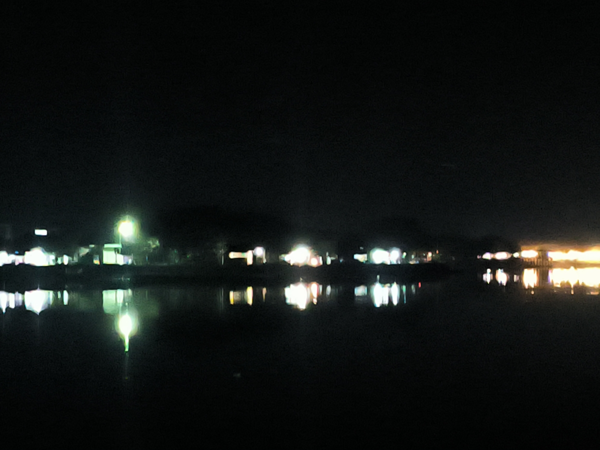 The image captures a nighttime scene of a waterfront area. Lights from buildings and structures are visible along the shore, with their reflections shimmering on the dark water. The location appears to be a settlement on the edge of a body of water, possibly in the city of Lhokseumawe, Indonesia. The darkness dominates the image, with the illuminated areas providing the primary points of interest.
