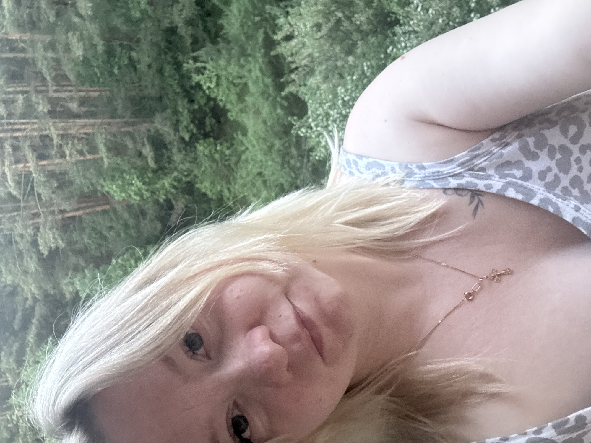 A woman with blonde hair is posing for a selfie in front of a forest. She is wearing a grey and white tank top and a delicate gold necklace. A tattoo is visible on her upper chest. The background shows a dense forest with tall trees. The location in the city of Chaikovsky, Russia is not ascertainable from the image.
