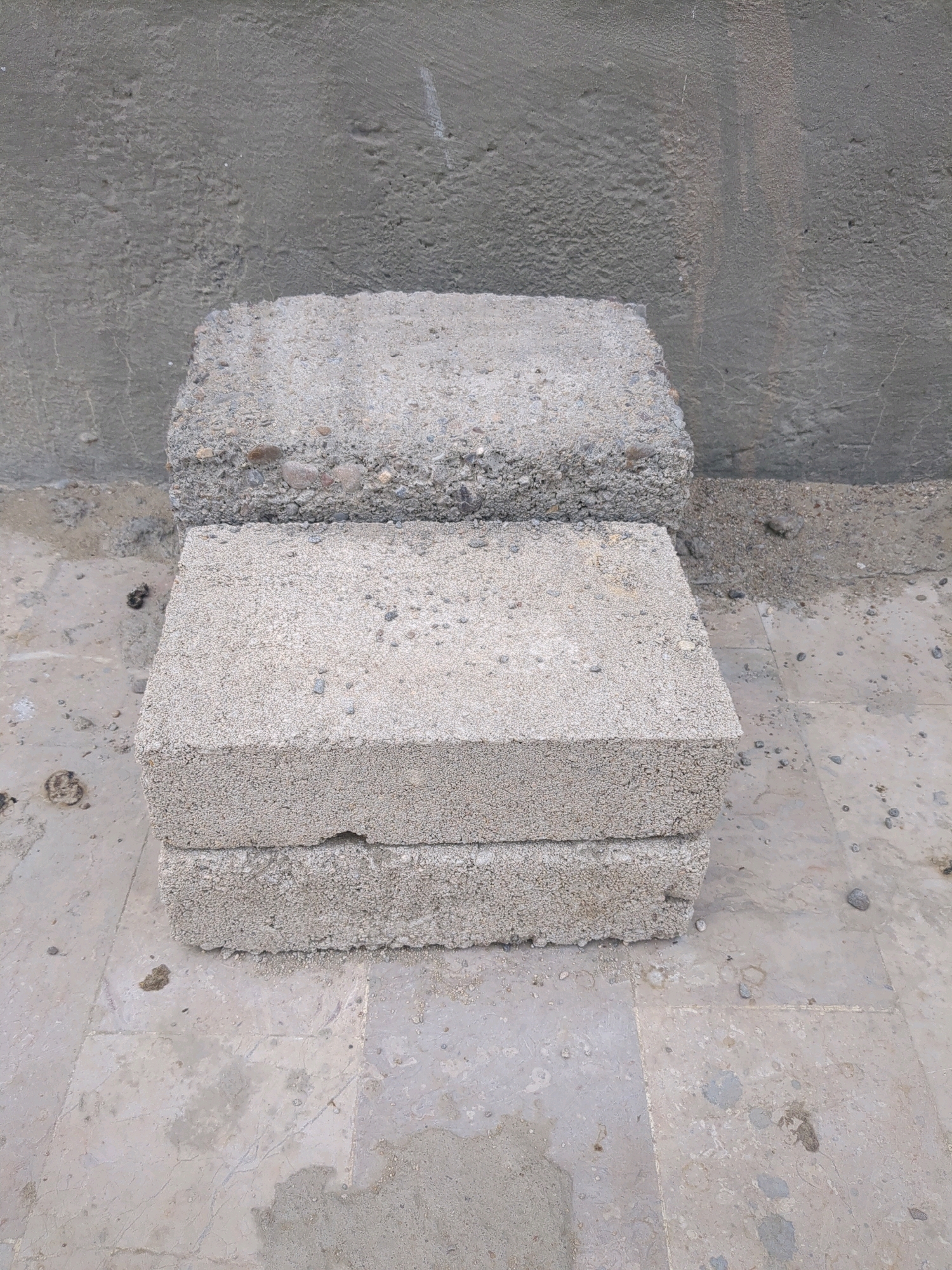Two concrete blocks are stacked on a tiled surface in front of a rough concrete wall. The blocks are light gray and appear to be of a standard size, with a porous texture. The lower block is partially obscured by the one above it. The tiled surface shows some wear and tear, with a few small pebbles scattered on it. The concrete wall in the background has a textured finish and is a darker shade of gray than the blocks. The lighting suggests an outdoor environment. The scene appears to be a construction or storage area. The image was taken in Karachi Division, Pakistan.