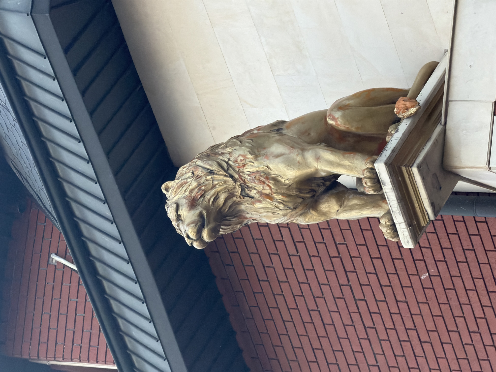 The image displays a large, ornate lion statue positioned as an architectural feature on a building, located in Sofia, Bulgaria. The statue, depicted in a rich golden or bronze hue with weathered accents of red and orange, shows the lion in a recumbent pose, its head resting on its forepaws. Intricate details are visible in its mane and musculature, suggesting skilled craftsmanship.
The lion rests on a light-colored plinth that appears to be integrated into the building's structure. Above the statue, a plain, light-toned wall or ceiling extends. Below the lion, a section of the building features a distinct pattern of red-brown bricks. To the left, a dark, ribbed structure, possibly a metal roof or awning, is visible, adding a contrasting modern architectural element. The lighting is even and bright, indicating it is daytime, likely under an overcast sky or in a shaded area, as no harsh shadows are cast. No activity is occurring, as the image captures a static facade detail. There is no visible text.