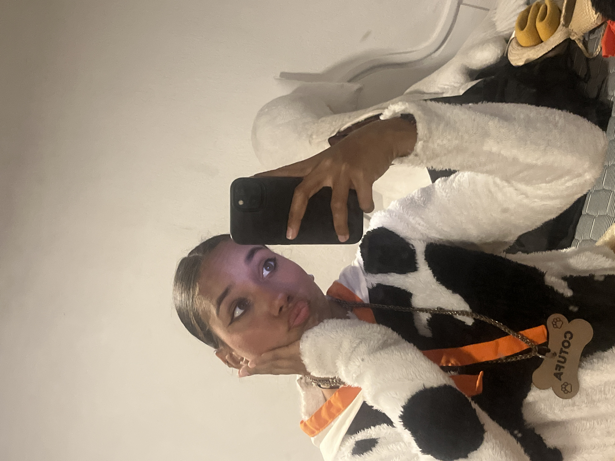 The image is a mirror selfie, rotated 90 degrees counter-clockwise, featuring a person dressed in a cow-themed costume. The subject, a young individual, has dark hair pulled back and is wearing eye makeup, including winged eyeliner. They are captured in a playful pose, with slightly pouted lips and resting their chin on their hands, looking upwards and slightly to the left.
They are wearing a fluffy, black and white cow-print top or costume, with an orange inner lining visible on the sleeve. A silver-colored chain necklace is visible, draped over the costume, featuring a light brown, bone-shaped pendant. The text "COUTUR" is visible on the pendant, appearing reversed due to the mirror reflection, along with two small paw print designs.
The person is holding a black smartphone with two camera lenses, which is used to capture the mirror selfie. The background primarily consists of a plain, light-colored wall. In the upper right portion of the image, some household items are visible, including a pair of yellow slipper-like shoes, a straw hat, and what appears to be a rack of clothes hangers or stored garments. The setting is indoors, with no specific time of day or weather cues. The overall impression is one of lightheartedness and playfulness.