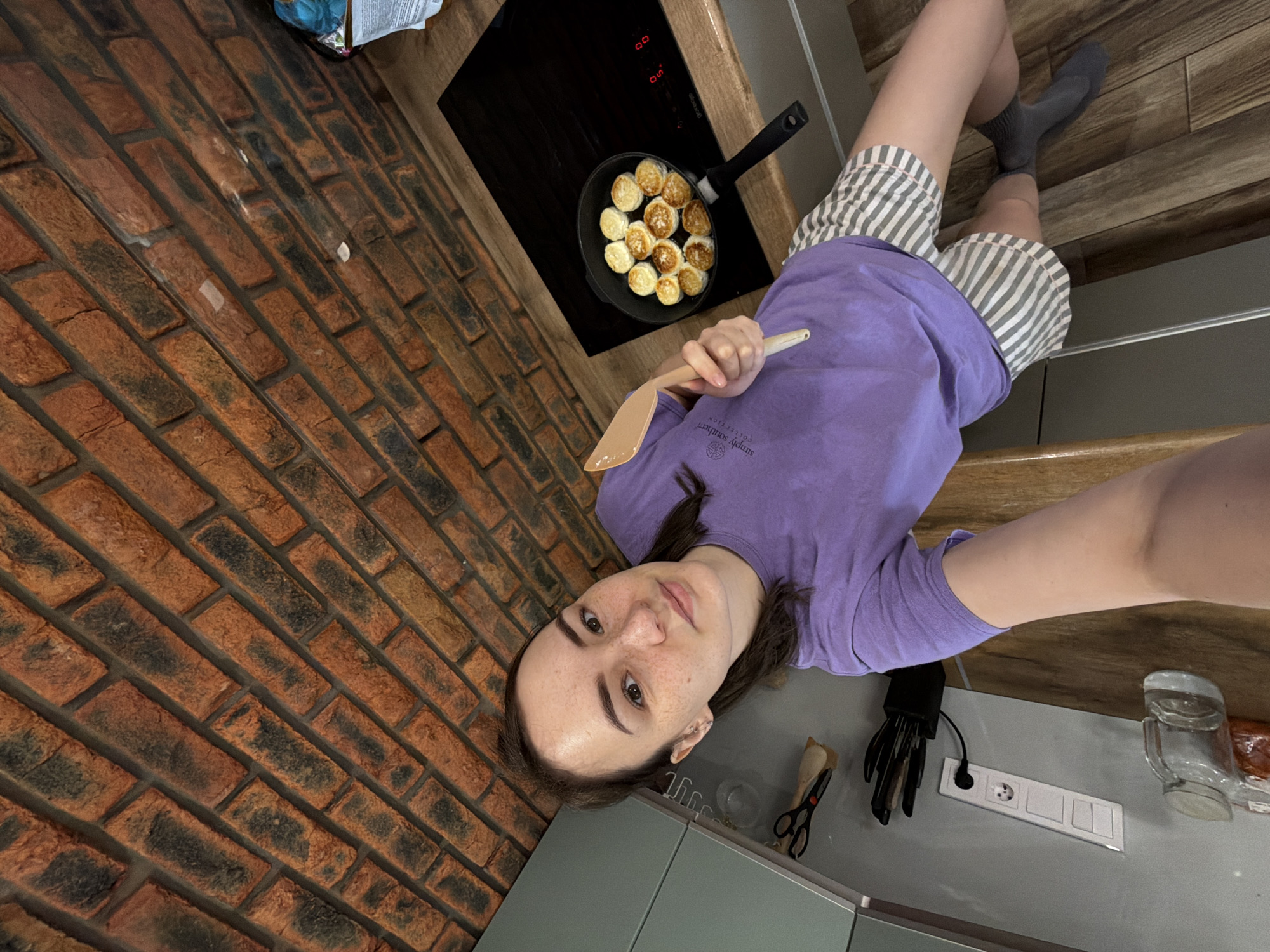 A person is taking a selfie indoors, presumably in a kitchen. The individual is wearing a lavender t-shirt and striped shorts. They are holding a wooden spatula. A frying pan filled with what appear to be small, round pancakes sits on a black stovetop in the background. The kitchen features a brick wall and modern grey cabinets. A power outlet with European plugs is visible on the countertop. The location is Chaikovsky, Russia.