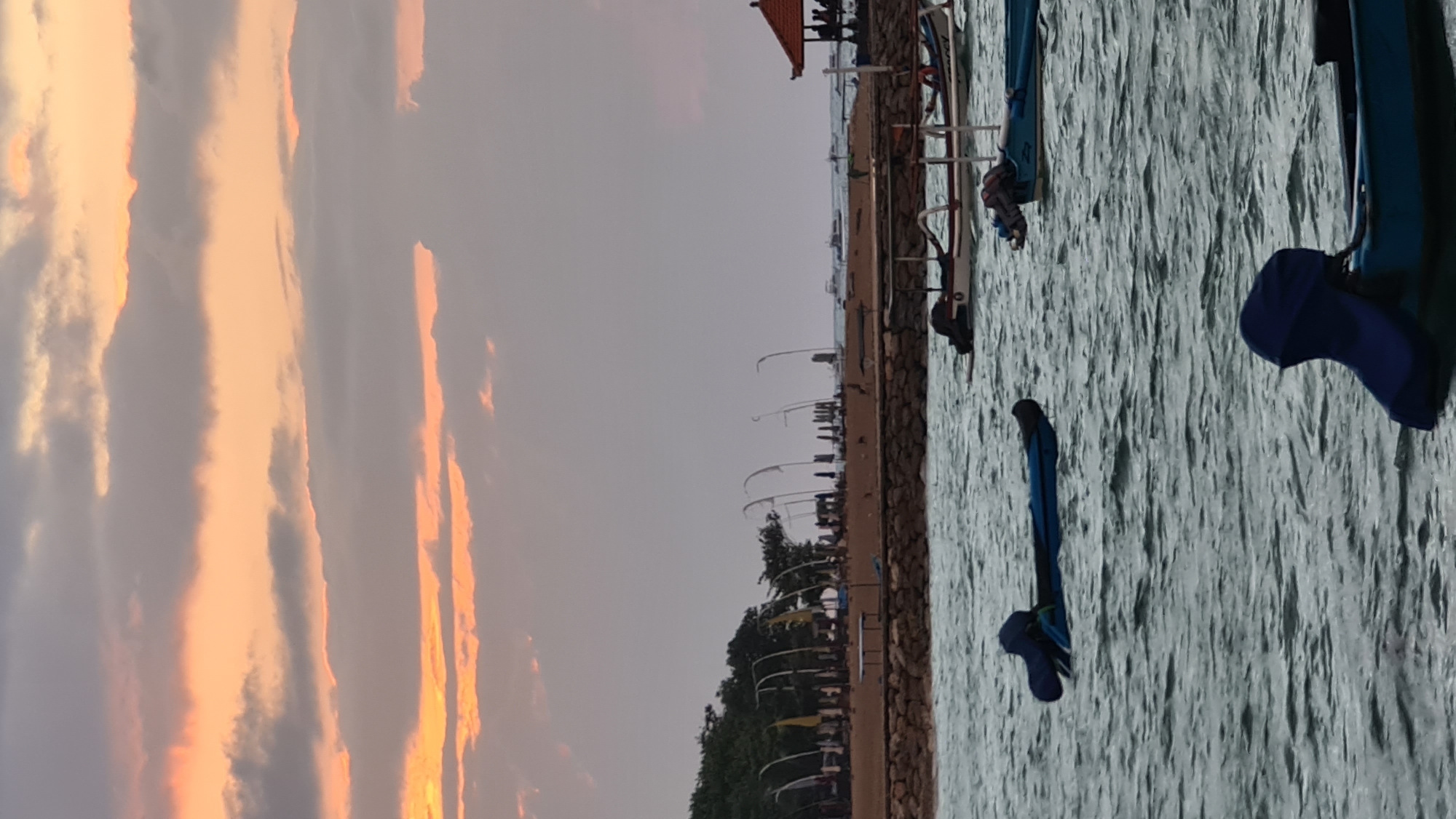 This is a vertically oriented photograph taken at sunset in Denpasar, Indonesia. The image depicts a tranquil scene with the sky painted in soft, warm hues of orange and pink from the setting sun, contrasting with the muted grey clouds. The foreground is dominated by the textured surface of the water, appearing choppy and reflecting the ambient light.
Along the edge of the water, a stone embankment separates it from a waterfront area. On this embankment, several boats are visible. Some are traditional outrigger canoes, characteristic of Indonesian waters. These boats are accompanied by various pieces of equipment, including what appear to be skis or surfboards and blue protective gear, suggesting recreational water activities. The presence of a small structure with an orange roof hints at a beachside establishment or a rental booth.
Further along the shore, a line of trees with lush green foliage creates a natural boundary. Beyond the trees, a series of curved structures, possibly part of a seating area or a recreational facility, are arranged along the waterfront. In the distance, a few indistinct figures of people can be seen, adding a sense of scale and human presence to the scene. The overall atmosphere is peaceful and serene, capturing the end of a day with a beautiful natural display.
