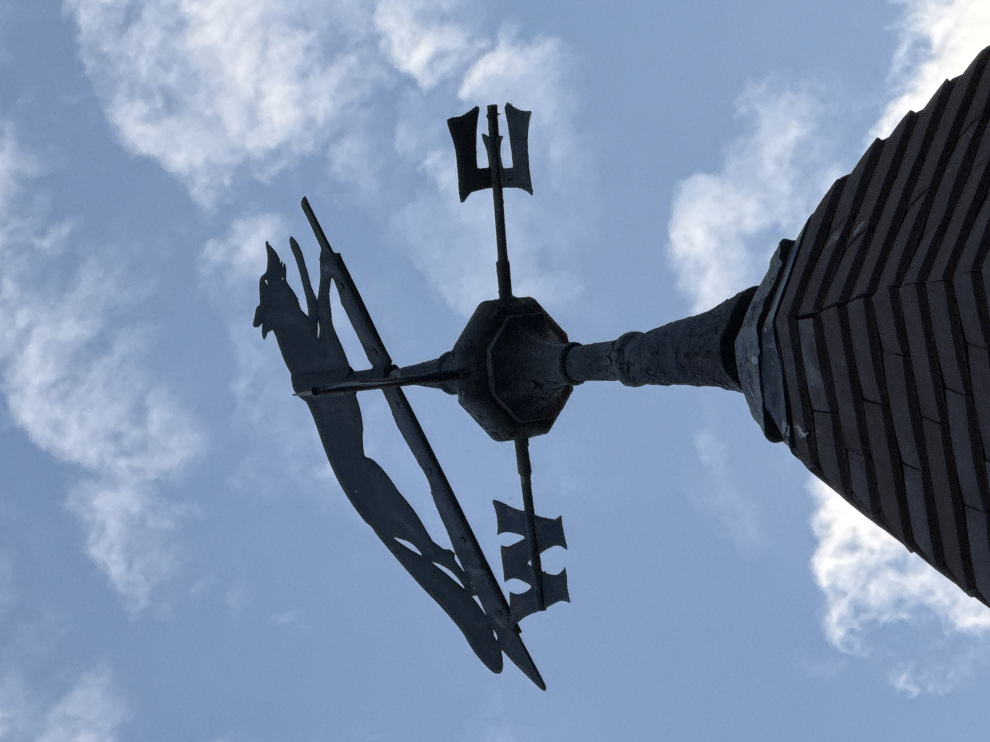 An upward shot reveals a weather vane mounted atop a building in London, UK. The vane is silhouetted against a sky with scattered clouds. The right side of the frame shows a portion of the building's roof, constructed with dark, overlapping materials, likely slate tiles. The weather vane itself has an elaborate design, including a central post with a spherical element and a direction indicator with ornate detailing. The tail of the vane is shaped like an animal, possibly a bird, with additional decorative elements. The overall scene has a historical or architectural character.