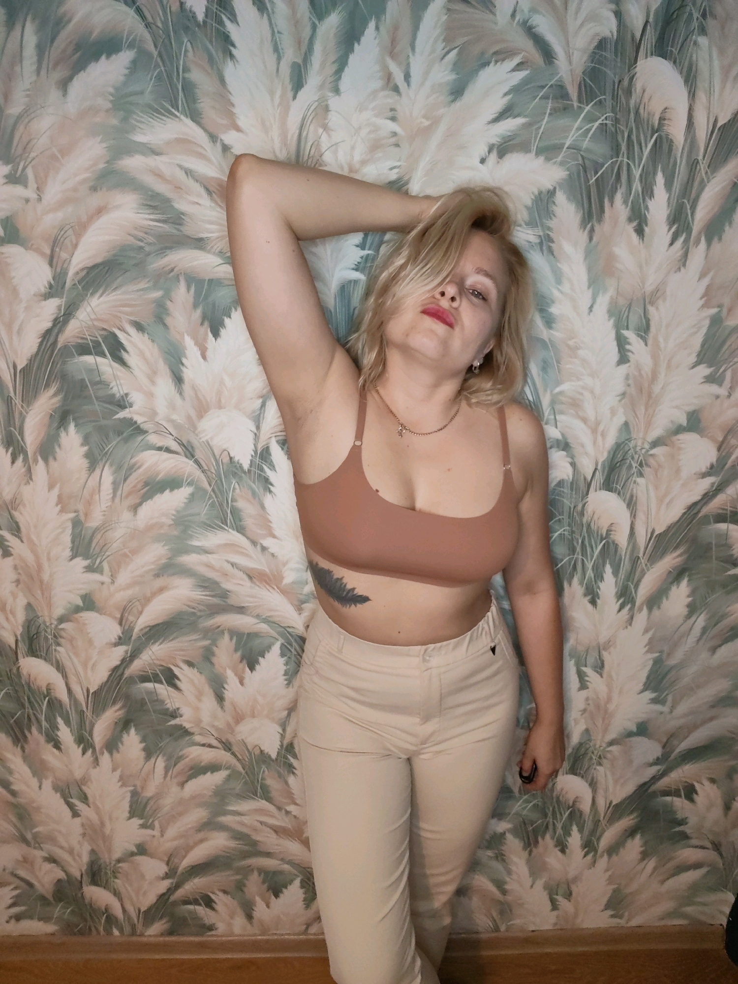 A blonde woman is posing in front of a wall with a beige and green floral pattern. She is wearing a tan bra and beige pants. Her left arm is raised above her head, and her hair is covering part of her face. She is wearing a necklace with a small cross pendant and earrings. There is a tattoo of a feather on her left side. She is also wearing a dark ring on her right hand. The floor is made of wood.