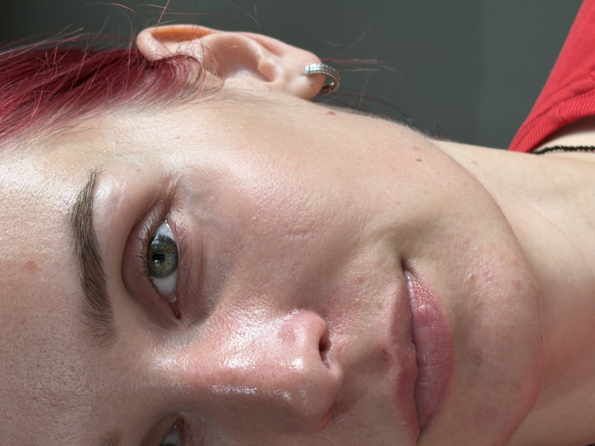 A close-up, chest-up, profile shot shows the right side of a person's face and neck. The person has fair skin, green eyes, dark eyebrows, and bright red hair. They are wearing a red shirt and a silver hoop earring on their right ear. The background is a plain dark gray. The image appears to be taken indoors, with the subject positioned in front of a plain, dark surface. There is no visible indication of the city of Chaikovsky, Russia, or any other specific geographic location.