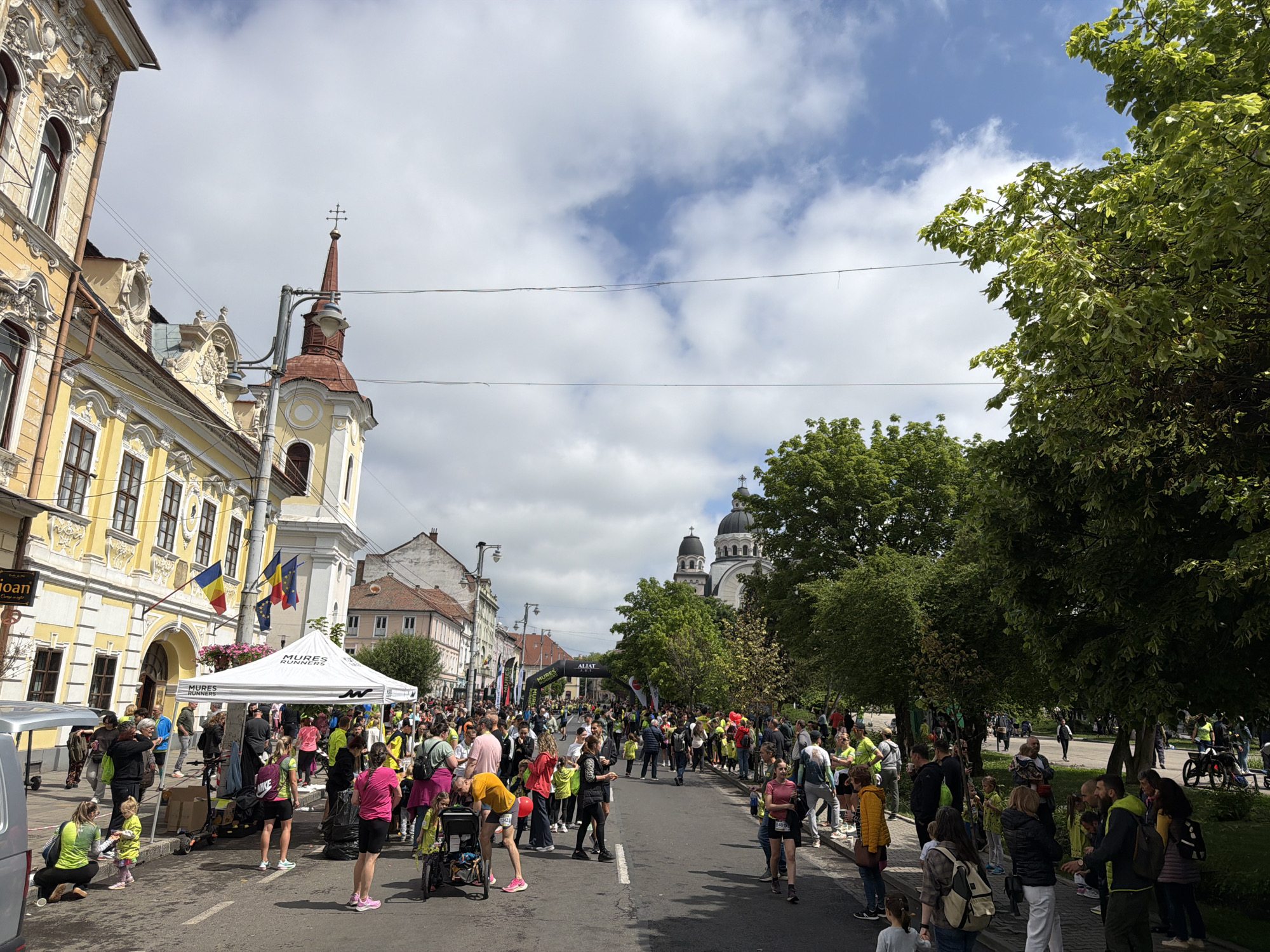 The image captures a street scene in Târgu Mureș, Romania, during what appears to be a public event, possibly a marathon or a race. A crowd of people is gathered along the street, with many wearing athletic attire. A banner or archway stretches across the road, marking the start or finish line of the event. A large tent with the inscription "Mures Runners" is set up on the left side of the image. On the left, a yellow baroque building with Romanian and European flags is prominent. In the background, a large cathedral with domes is visible. The sky is partly cloudy with patches of blue. Trees line the right side of the street, and various vehicles, including vans, are parked nearby. People are engaged in various activities such as walking, stretching, and socializing.