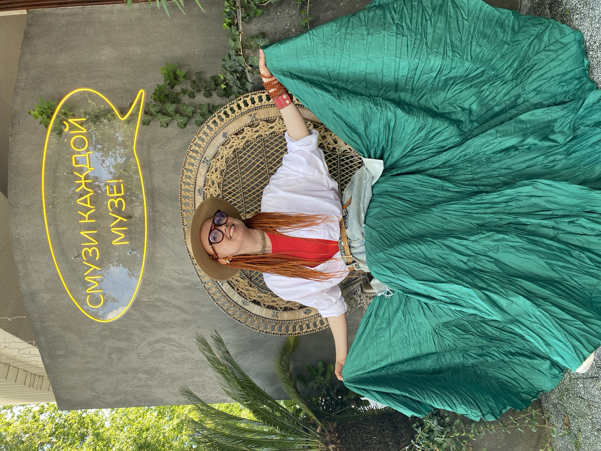 In Sochi, Russia, a young woman with vibrant orange braided hair and glasses sits on a large wicker chair outdoors. She is wearing a straw hat, a red top, a white open shirt, denim shorts, and a voluminous, pleated green skirt that drapes around her. She is smiling and looking to her right. Behind her, a concrete wall features a yellow neon sign in the shape of a speech bubble that reads "СМУЗИ КАЖДОЙ МУЗЕ!" which translates to "Smoothies for Every Muse!". Lush green plants, including a palm tree, grow around the scene, and the lighting suggests it is daytime.