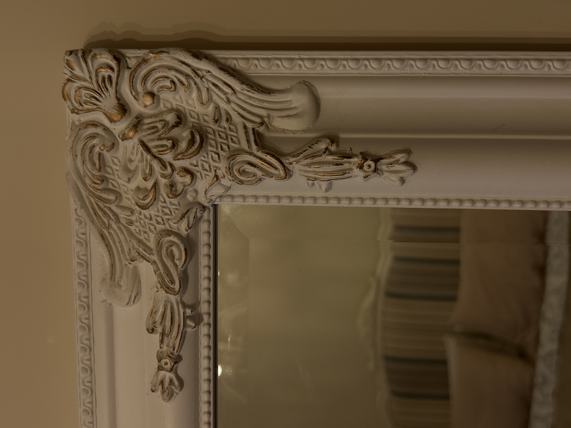 The image shows a close-up of the ornate, white and gold corner of a mirror frame. The frame features intricate carvings and moldings. The mirror reflects a blurred interior, potentially showing a striped pattern and a light-colored object. The background wall is a light beige color. This image does not provide information to identify a location in Plovdiv, Bulgaria.