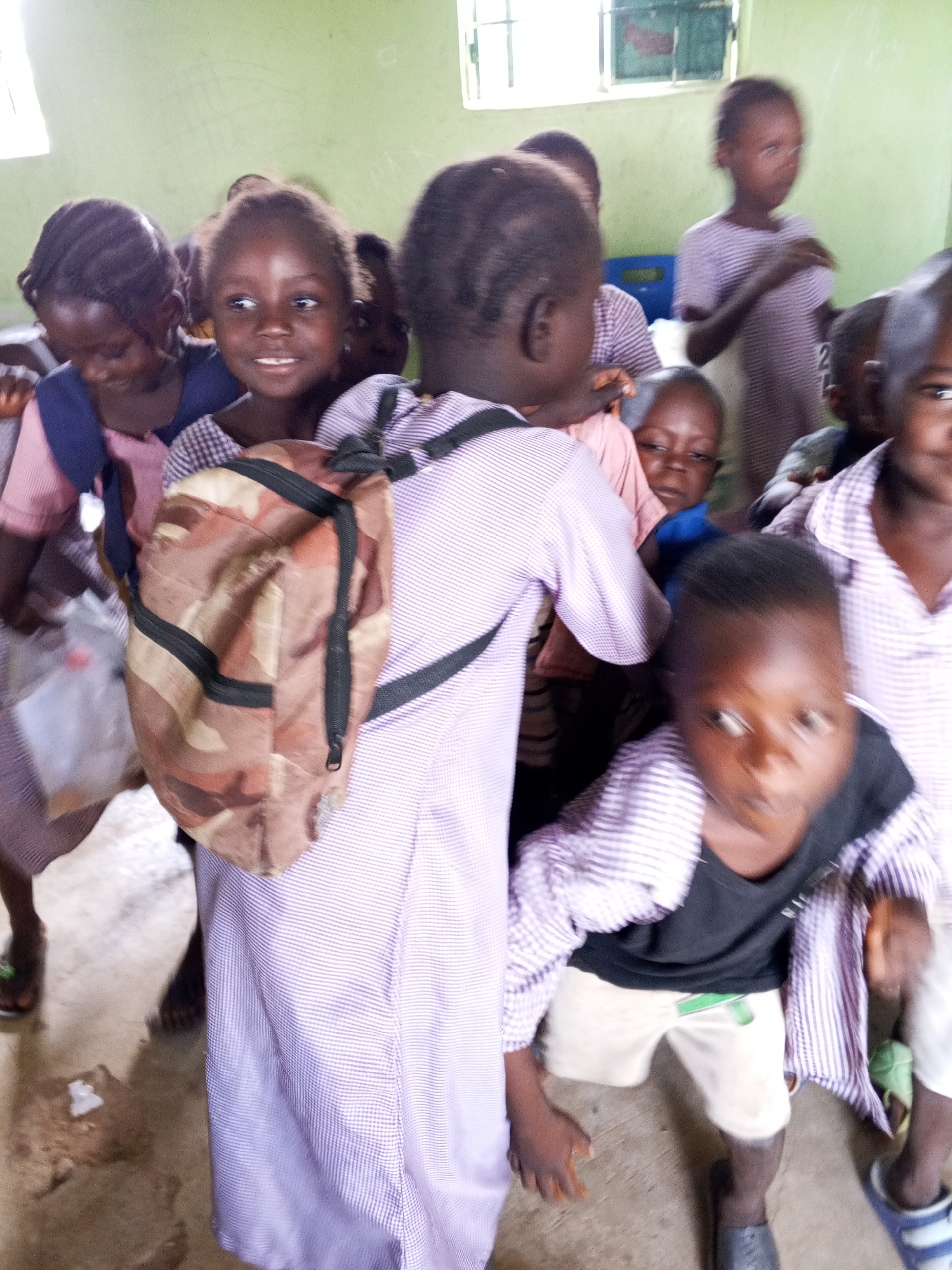 The image depicts a group of young children, appearing to be students, in a classroom setting. The room has light green walls and a window with visible bars. Some children are wearing uniforms, characterized by light purple and white striped shirts. One child in the foreground is wearing a camouflage backpack. The floor appears to be concrete and shows signs of wear and some moisture. The children are gathered closely together, suggesting a communal activity or a crowded space. The scene is likely in Andaha, Nigeria, given the context provided, and suggests an educational environment for young students.
