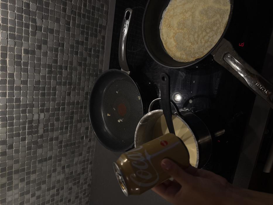The image displays a kitchen setting with a focus on an induction cooktop and a tiled backsplash. A human left hand holds an open, golden-colored Coca-Cola can over a metal saucepan containing a light-colored, viscous liquid. A black stirring spoon rests within this saucepan. On the black induction cooktop, which bears a "BOSCH" logo and displays the digit "6," two non-stick frying pans are visible. One pan, located in the foreground to the right, contains a single, cooked pancake or crepe. The second pan, positioned further back and to the left, is empty save for minor residue on its cooking surface. The backsplash consists of small, square, dark gray tiles. No discernible visual markers within the image provide evidence to confirm the requested location in the city of Warsaw, Poland.