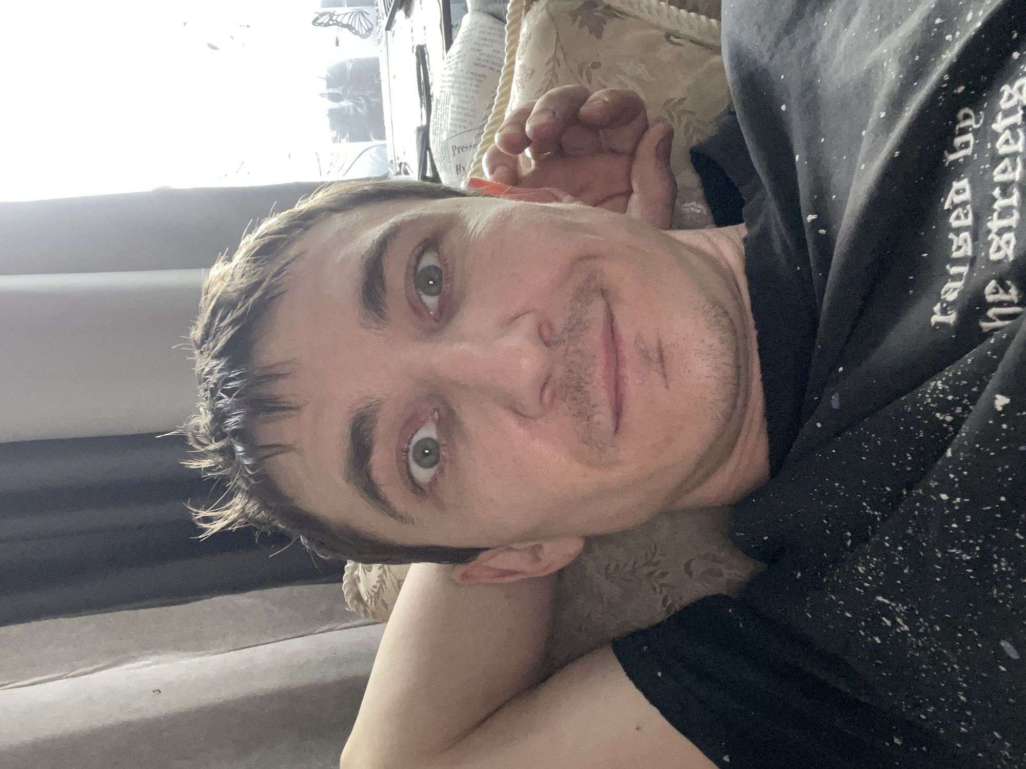 A man is lying on his back on a couch, with his head resting on a pillow. He is looking at the camera with his eyes wide open. His right arm is behind his head, and his left hand is resting on the pillow. The man is wearing a black t-shirt with white splatters on it. The background is a blur of curtains and a window. The scene is indoors, likely in a residential setting. There is no indication of the location in Laishevo, Russia.