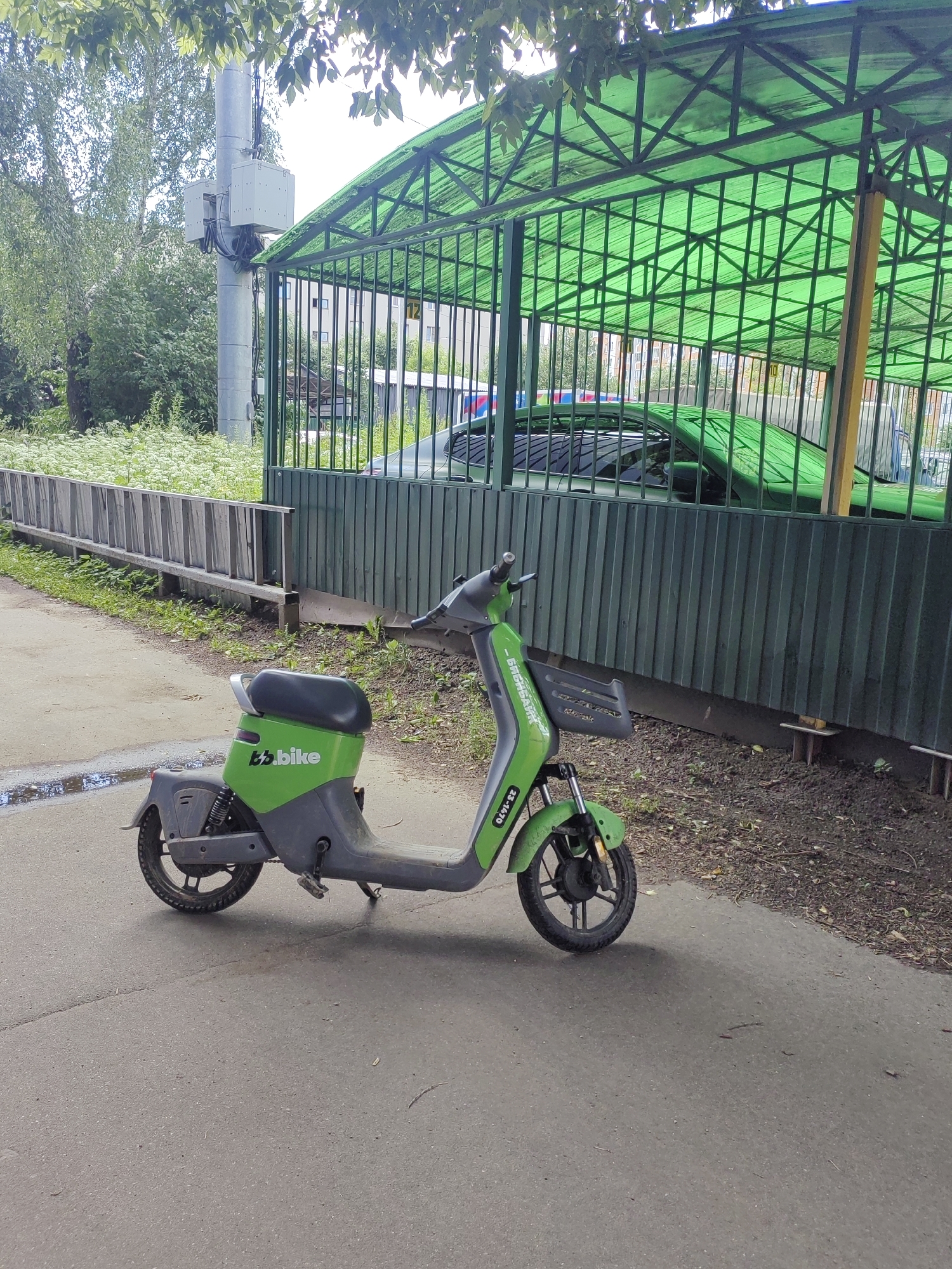 A green and grey electric scooter, labeled "B.bike", is parked on an asphalt path next to a wooden fence. Behind the fence is a green metal canopy with a transparent roof, under which a dark green car is parked. The car's roof and hood are visible. The area behind the canopy appears to be a residential building with windows. Trees and foliage are present in the background, and the sky is overcast. The scooter has a kickstand extended. A puddle of water is visible on the asphalt near the scooter. The scooter has a license plate or identification number on its front fender, reading "23-1470". The scene is likely in Putilkovo, Russia, based on the context provided.