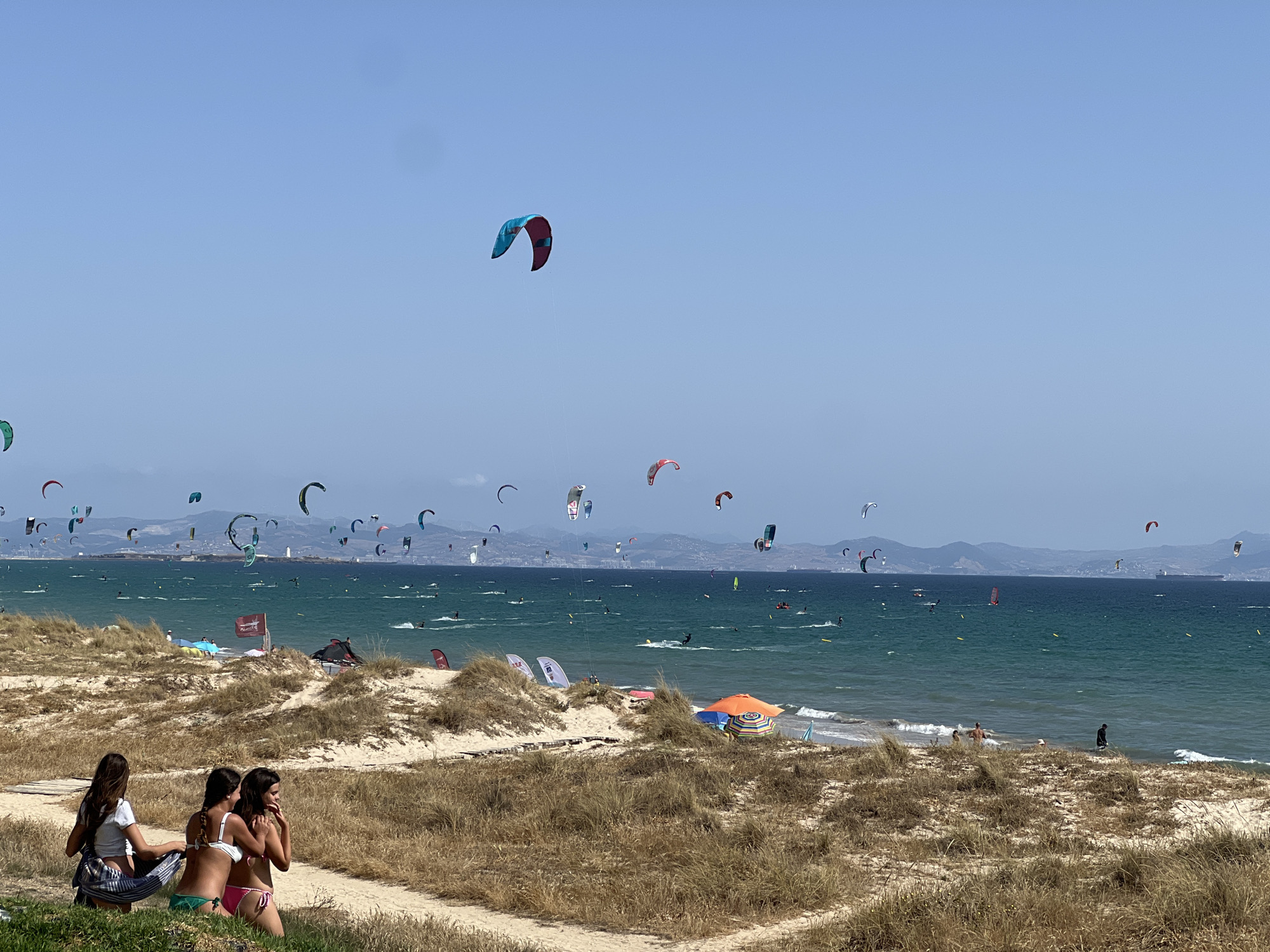 The image depicts a sunny day at a crowded beach in Tarifa, Spain, known for its windy conditions and popularity among kitesurfers. In the foreground, three young women are seated on a grassy embankment overlooking the sandy beach and the ocean. They are dressed in bikinis and appear to be relaxing and socializing. Behind them, a vast expanse of turquoise water is filled with numerous kitesurfers, their colorful kites dotting the sky. The activity on the water suggests a dynamic and energetic atmosphere. The background shows a hazy coastline with mountains, indicating the presence of land beyond the sea. The overall scene captures a vibrant beach environment characterized by water sports and leisure activities.