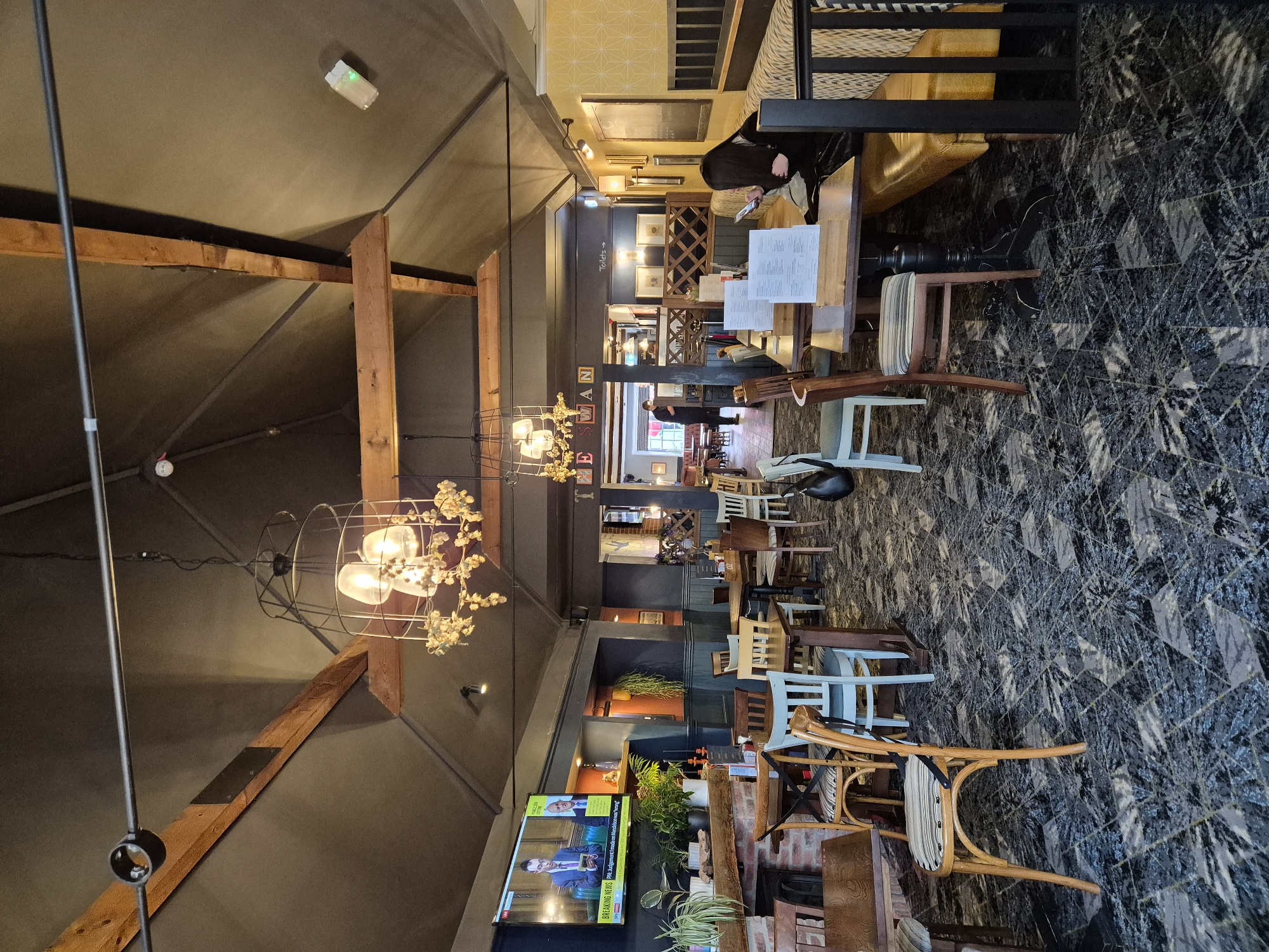 This is an interior wide-angle shot of a pub or restaurant, characterized by a high, vaulted ceiling with exposed wooden beams and a dark, possibly fabric-covered, finish. The space is furnished with various seating arrangements, including a long booth with yellow upholstered benches along the back wall and numerous tables with a mix of wooden, upholstered, and light blue chairs scattered across a dark, patterned carpet.
Several light fixtures hang from the ceiling, including large, industrial-style chandeliers adorned with faux flowers. Artificial lighting is prominent, supplementing natural light, though no direct windows are clearly visible.
In the background, a person with dark hair is seated at a table in the booth area, looking at a mobile phone, with several menus displayed on the table. Another individual is partially visible further back near what appears to be a bar or service area. On the left side of the frame, a large flat-screen television is mounted on a wall, displaying a news program featuring two men, with a news ticker reading: "BREAKING NEWS PM-Agement I made on Mandelson was wrong". Below the TV are potted plants and a brick-faced structure, possibly a fireplace.
Visible text includes directional signage for "Toilets →" and a partial establishment name, "THE SMAN". The setting suggests a casual dining or social venue.