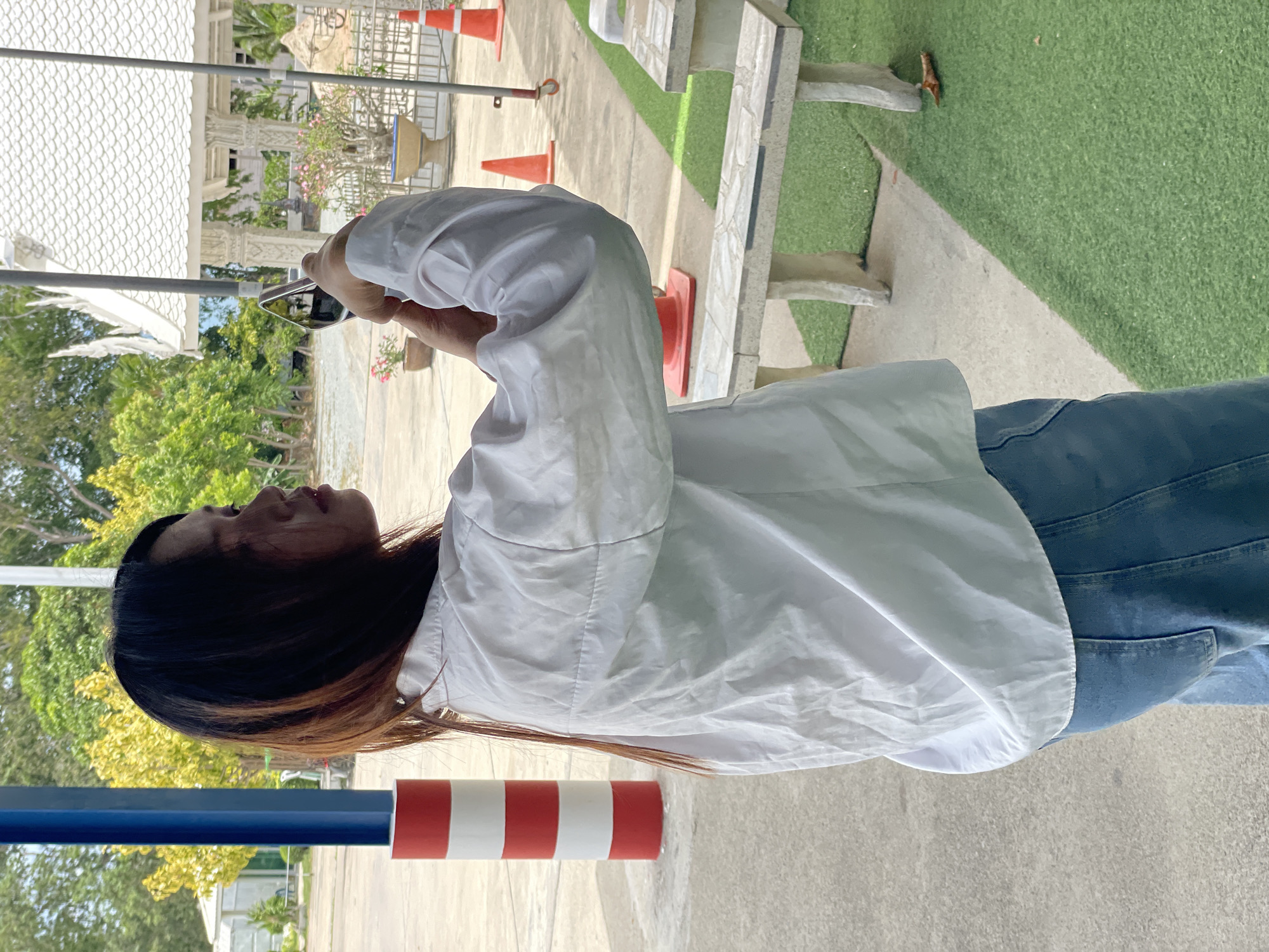In Tambon Na Chom Thian, Thailand, a young woman with long, dark hair, partially lightened at the ends, is shown from the back, facing to her right and looking into a smartphone she holds with both hands. She is wearing a loose-fitting white button-up shirt and blue denim jeans. The background features a paved area, a bright green artificial turf, and a white, ornate building with a tiled roof, characteristic of a Thai temple. A striped red and white pole stands to the left of the woman, and orange traffic cones are visible in the distance. The scene appears to be outdoors during daylight.