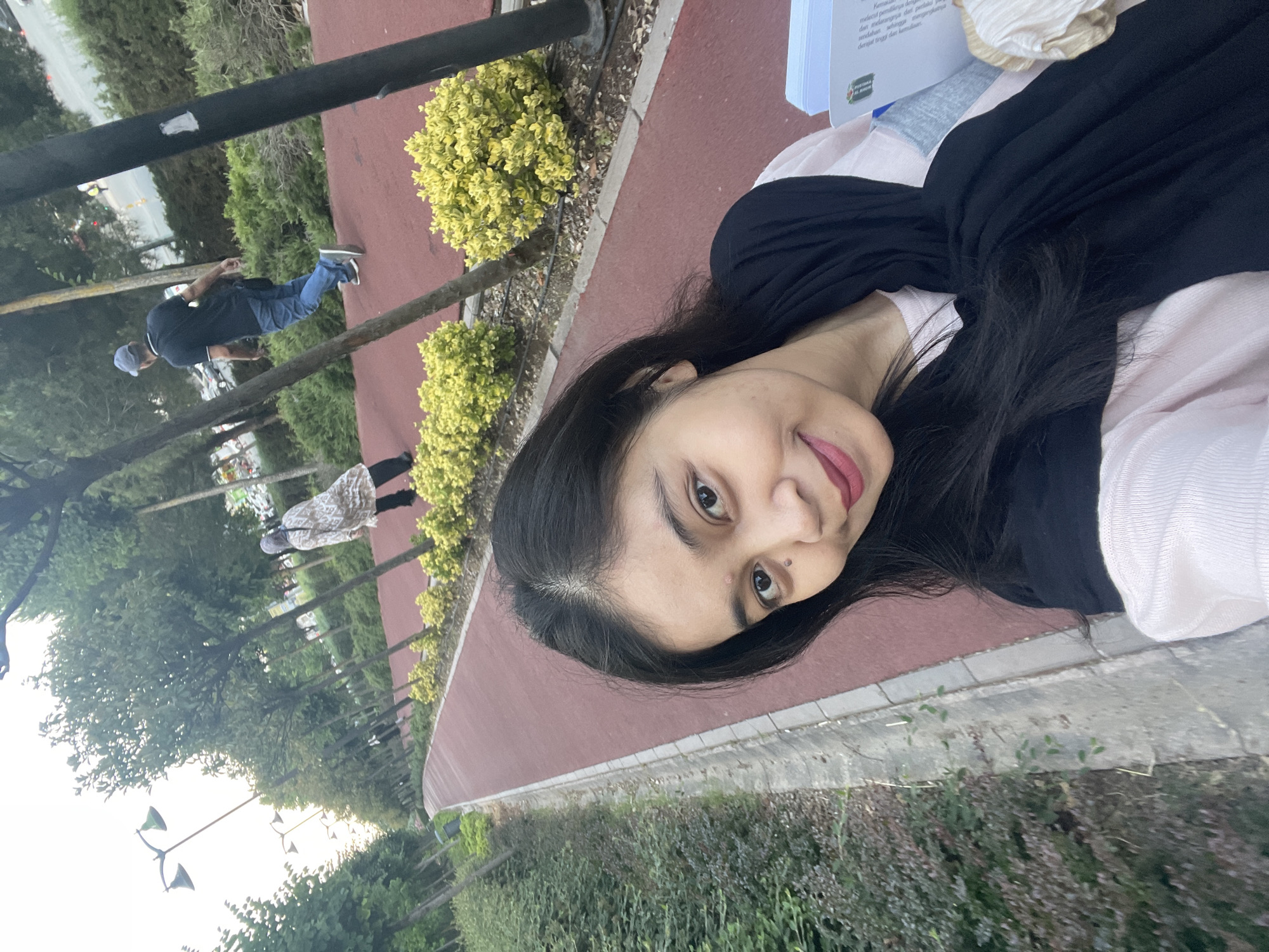 A young woman with long, dark hair and light skin takes a selfie in a park in Başakşehir, Turkey. She is wearing a light pink top and a black scarf. In the background, a red walking path curves through a park with trees and yellow bushes. Two other people are visible in the distance: one person wearing a patterned dress and a headscarf, and another person in a dark blue shirt and jeans, walking away from the camera. Streetlights are visible along the path. The woman is holding a book or folder in her right hand.