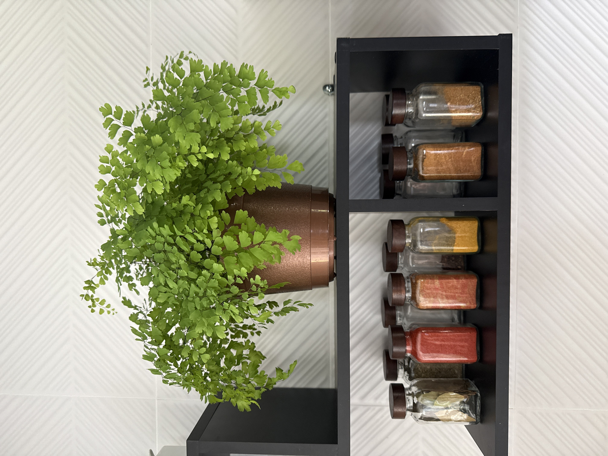 A fern in a copper pot sits on a black shelf, above a collection of spice jars. The shelf is mounted on a white tiled wall. The spice jars are clear glass with dark brown lids and contain various spices including red paprika, yellow turmeric, and other ground spices. The wall tiles have a subtle, diagonal pattern.