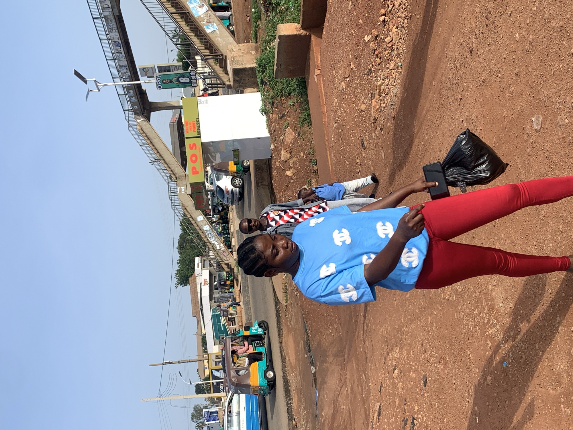 The image depicts a street scene in Gura Top, Nigeria. A young woman in a light blue t-shirt with a repeating pattern of interlocking 'C' shapes and red leggings is the primary subject, holding a black mobile phone and a black plastic bag. To her right, another person in a patterned shirt and grey pants is visible, and further back, a man in a blue shirt is lying down or resting. The background shows a dusty, reddish-brown road with scattered stones.
In the middle ground, there is an elevated walkway or bridge structure with railings, supporting advertisements and a street light. Buildings, including one with a large "POS" sign, are visible below the walkway. Various vehicles, including a white car, a yellow and teal auto-rickshaw with passengers, and a blue bus, are present on the road. Lush green vegetation can be seen in the distance.
The lighting suggests it is daytime, likely late morning or early afternoon, with clear skies. The overall atmosphere appears casual and possibly indicative of a bustling commercial or transportation hub. The presence of auto-rickshaws and the general street environment are characteristic of Nigerian urban or semi-urban areas. Some text is visible on the advertisement boards, including "POS" and "OPay".