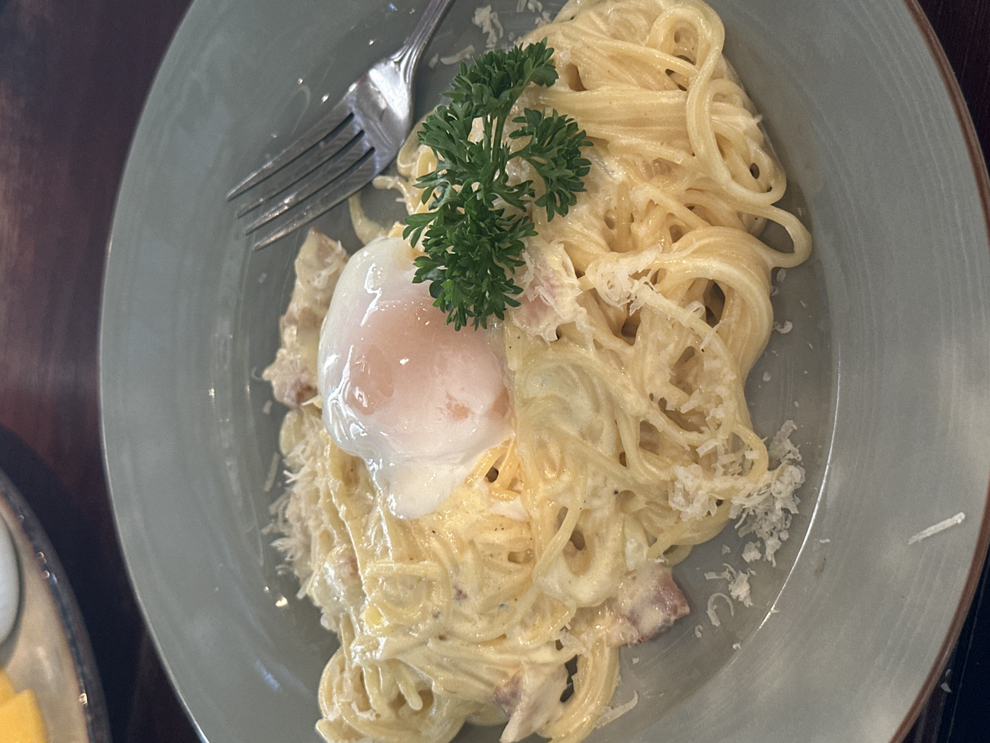 A high-angle, close-up shot shows a plate of spaghetti carbonara, garnished with a poached egg and a sprig of parsley, sitting on a table. A fork rests near the egg. The plate is light grey-green and has a slightly uneven, handmade appearance. The spaghetti is coated in a creamy sauce, with visible pieces of bacon and grated cheese. The poached egg has a runny yolk. The table appears to be made of dark wood. This image was taken in Pattaya City, Thailand.
