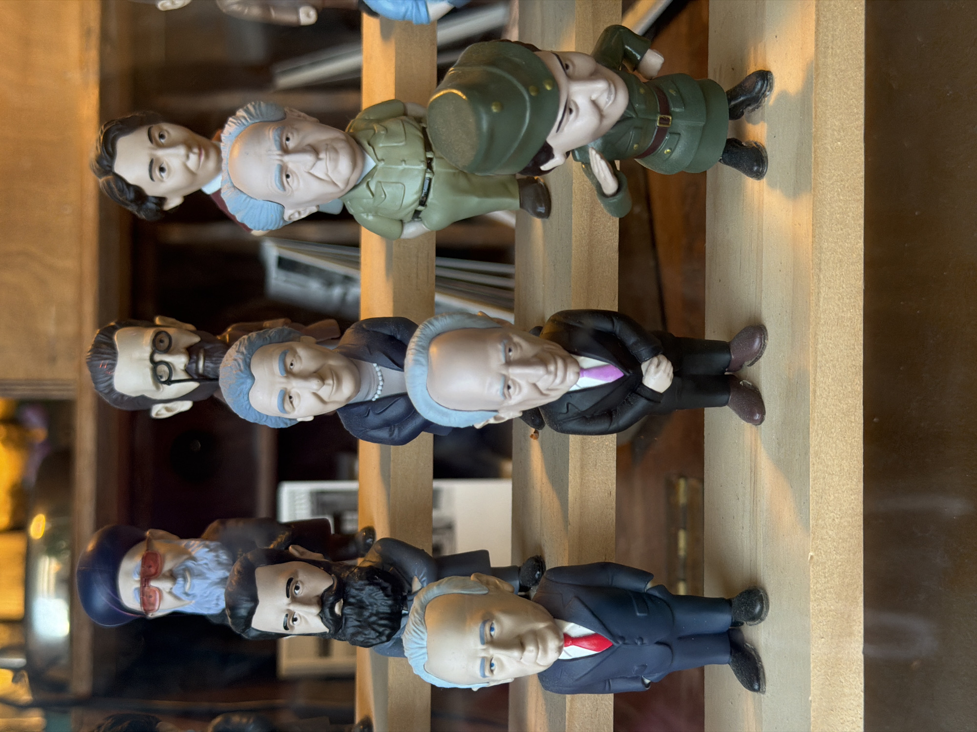 The image shows a collection of bobblehead figures displayed on wooden shelves in what appears to be a store or a display case. The figures are arranged in rows and depict various individuals, many of whom are recognizable public figures, likely politicians or historical personalities.
In the front row, from left to right, are two figures in suits. The leftmost figure wears a dark suit, a white shirt, and a red tie, with hands tucked into his pockets. To his right is another figure in a dark suit, white shirt, and pink tie, with hands clasped in front. Further to the right, a figure in a green military uniform with a hat is shown saluting. This figure has short brown hair and is wearing a belt and boots.
The middle row features several figures. On the left, a figure in a dark suit and red tie stands with hands in pockets. Next to him, a figure with a blue shirt and dark jacket with a pearl necklace is visible. To the right of this, a figure in a light green military-style uniform with a belt is standing with hands in pockets.
The back row shows more figures, including one with a dark blue religious hat and red-tinted glasses, a long blue beard, and a dark coat. Next to him is a figure wearing glasses and a dark beard. To the right of these, a figure with dark hair and a light-colored shirt is positioned behind a figure with light blue hair and a green shirt.
The background is blurred, but it suggests a shelving unit with books or other items, illuminated by warm lighting, creating a soft glow. The overall impression is that of a curated display of collectible figures. The image itself was captured without any specific indication of its location in Tel Aviv, Israel.