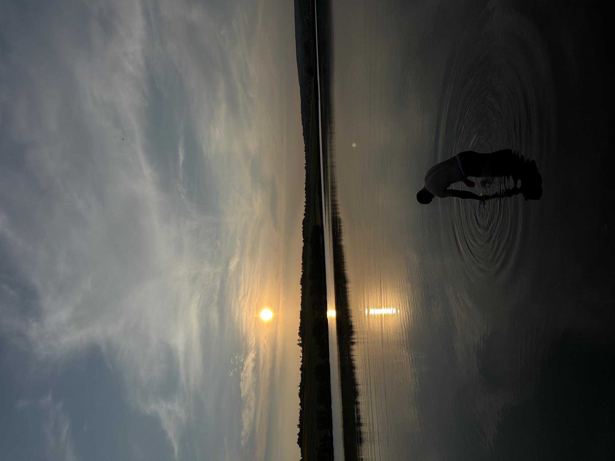 The image captures a serene sunset over a tranquil lake in Găleștii Noi, Moldova. A lone figure, a shirtless person wearing dark shorts, stands knee-deep in the water, creating ripples with their hand. The sun, a bright orb, is low on the horizon, casting a golden glow across the sky and reflecting on the water's surface. The landscape is dotted with trees and rolling hills in the distance. The sky is a canvas of soft clouds, tinged with hues of blue and orange.