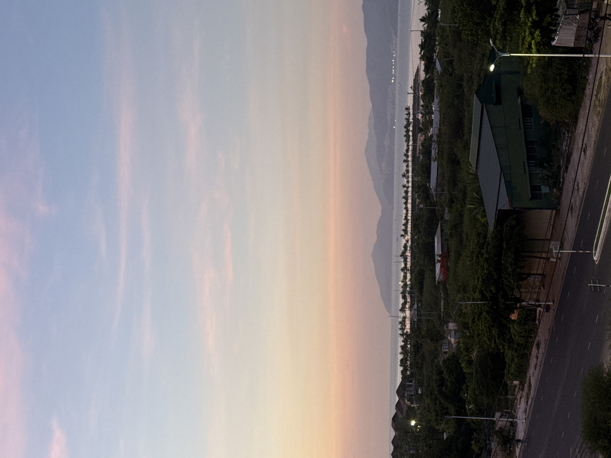 The image depicts a coastal landscape at dusk in Bac Nha Trang Ward, Vietnam. The sky transitions from pale blue at the top to soft pink and orange hues closer to the horizon, with thin, wispy clouds. In the distance, a line of mountains forms the backdrop to the calm sea, dotted with lights that suggest a populated coastline. Along the shore, a dense row of palm trees stretches horizontally, separating the water from the urban fringe.
In the foreground, a road runs diagonally across the lower part of the frame. Adjacent to the road, there are buildings and dense vegetation. One prominent building, a dark green, angular structure with a metal roof, stands out amongst the greenery. Streetlights illuminate parts of the scene, casting a yellow glow. There are no discernible people in the frame, and the overall atmosphere is one of quietude as day transitions into night.