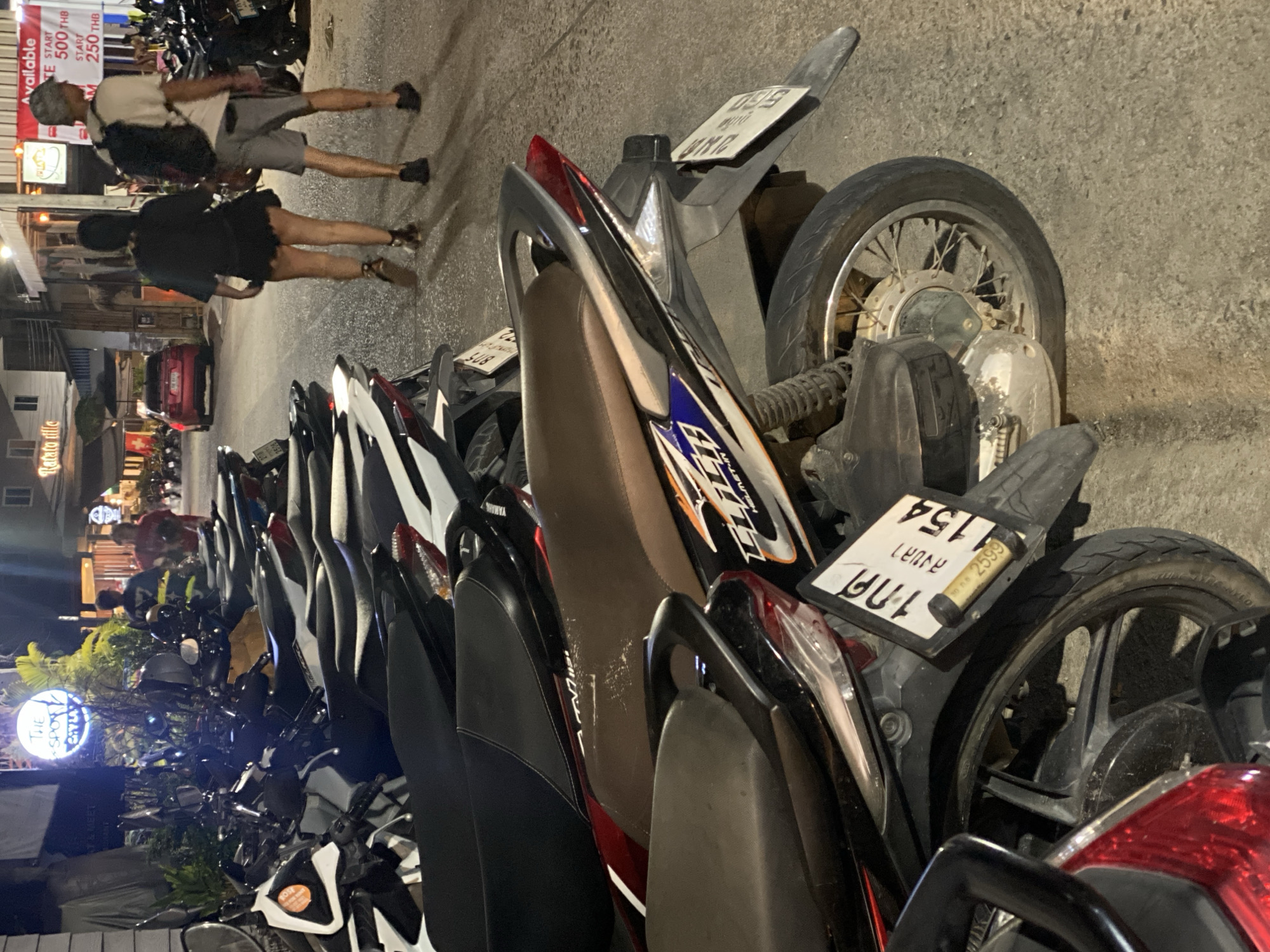 The image depicts a row of parked motorbikes along a street in Baan Kai, Thailand. A man and a woman are walking away from the camera, in the background. A red car is parked by the side of the road. The motorbikes are tightly packed together, with varying colors. A round lit sign with a tree on it reading "The Spot Gyla" is visible, as is a shop sign with text on it, partially obscured by the couple. A sign listing prices in Thai Baht (THB) is visible.