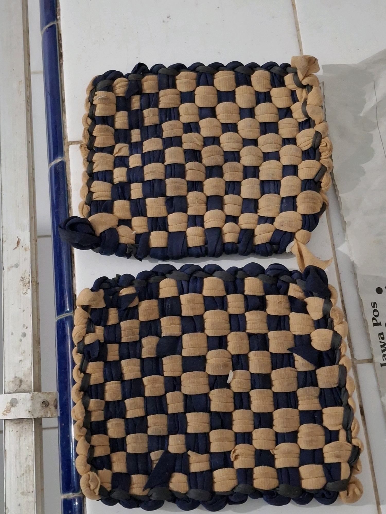 Two woven potholders, one atop the other, are displayed against a tiled wall. The potholders exhibit a checkerboard pattern created by interweaving strips of dark blue and tan fabric. The tan fabric appears slightly worn and frayed. The background wall consists of white tiles with some visible grout lines. To the left of the potholders, a blue tiled column runs vertically, and a metal railing is partially visible. To the right, a section of white paper with faint black text is present. The overall setting suggests an indoor environment, possibly a kitchen or storage area in Lumajang, Indonesia.