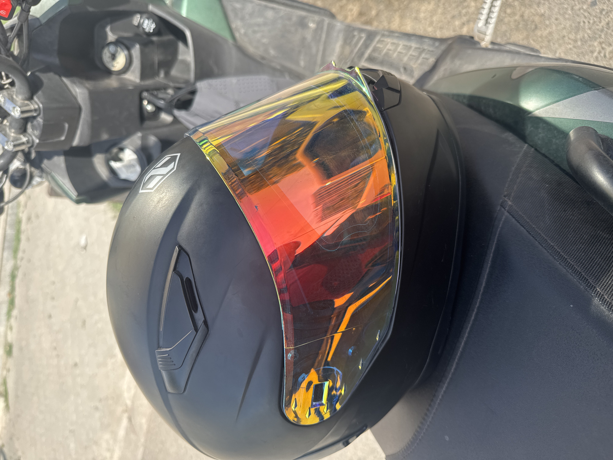 A matte black motorcycle helmet with a reflective, multi-colored visor rests on the dark gray seat of a green and black scooter. The scooter's handlebars and part of its front are visible in the background, along with a concrete surface and some out-of-focus vegetation. The scene appears to be outdoors, possibly in Bishkek City, Kyrgyzstan, given the context. The lighting suggests it is daytime.